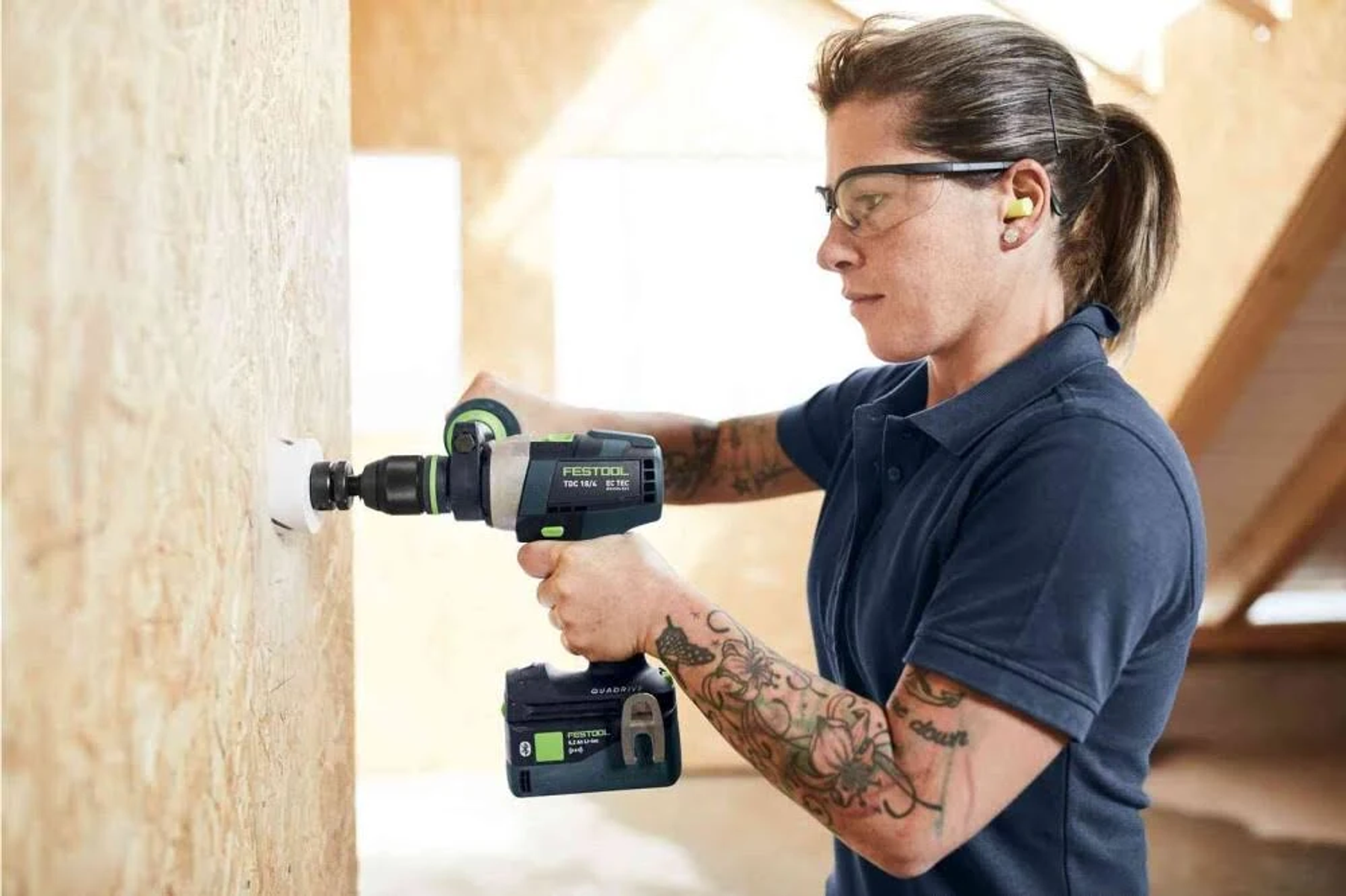 Una mujer taladrando un agujero en una pared de madera con un taladro Festool, usando gafas de seguridad.