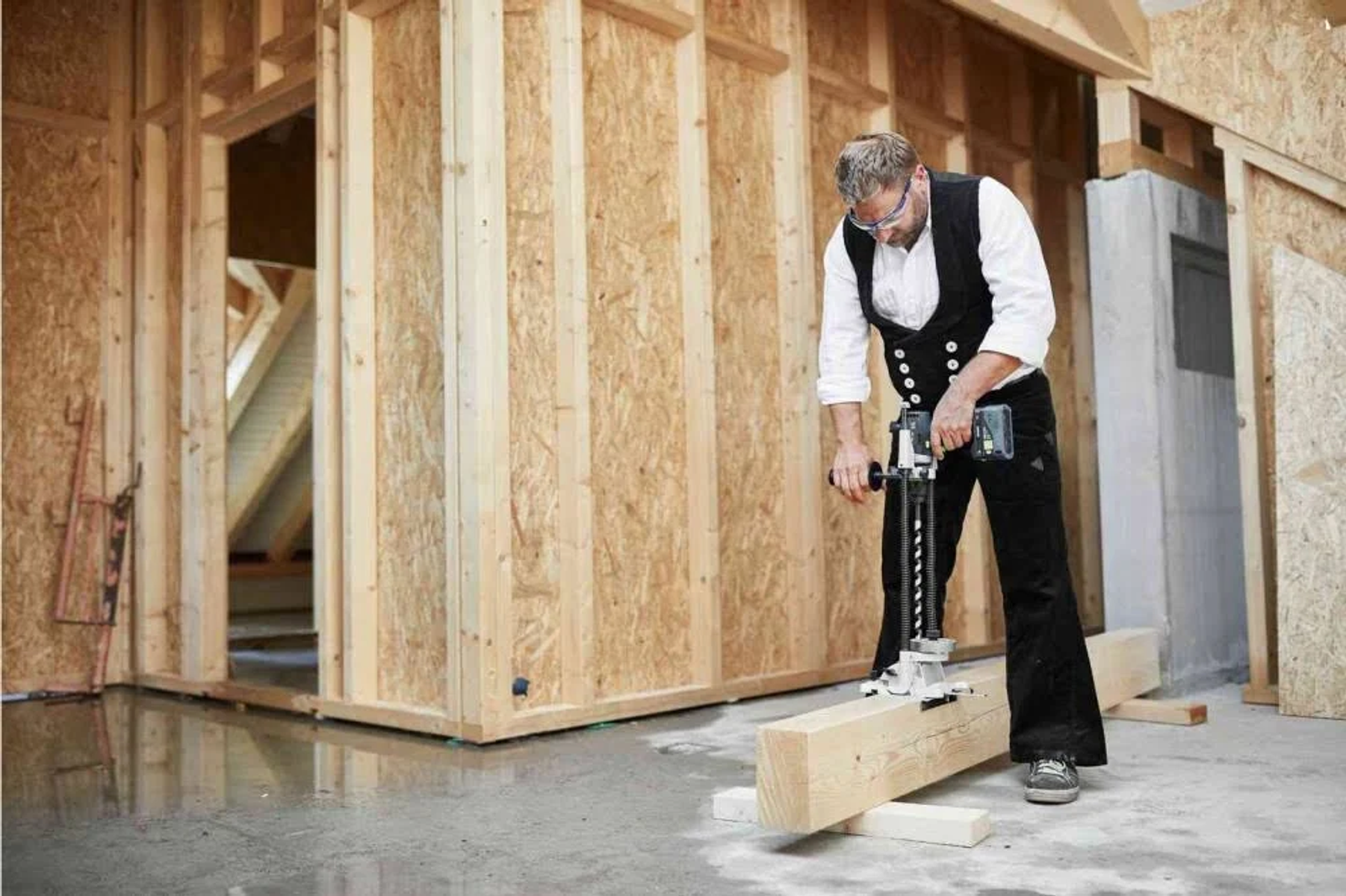 Un hombre taladrando una viga de madera, vistiendo ropa de trabajo, dentro de un edificio con estructura de madera en construcción.