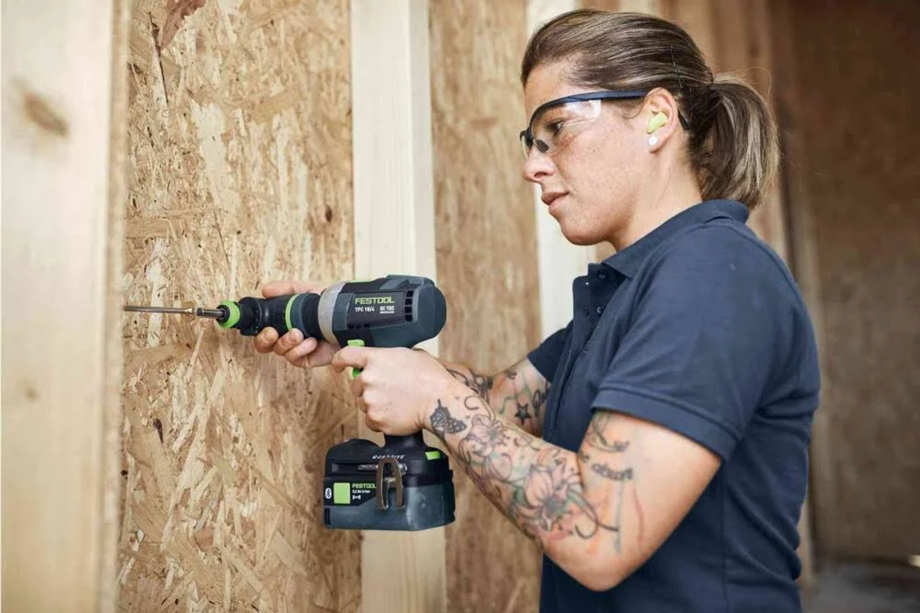 Una mujer con gafas de seguridad taladra madera con un taladro eléctrico verde y negro.