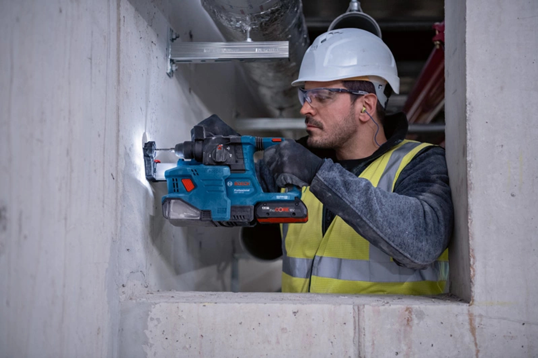 Hombre usando un taladro azul para perforar una pared blanca, usando equipo de seguridad.