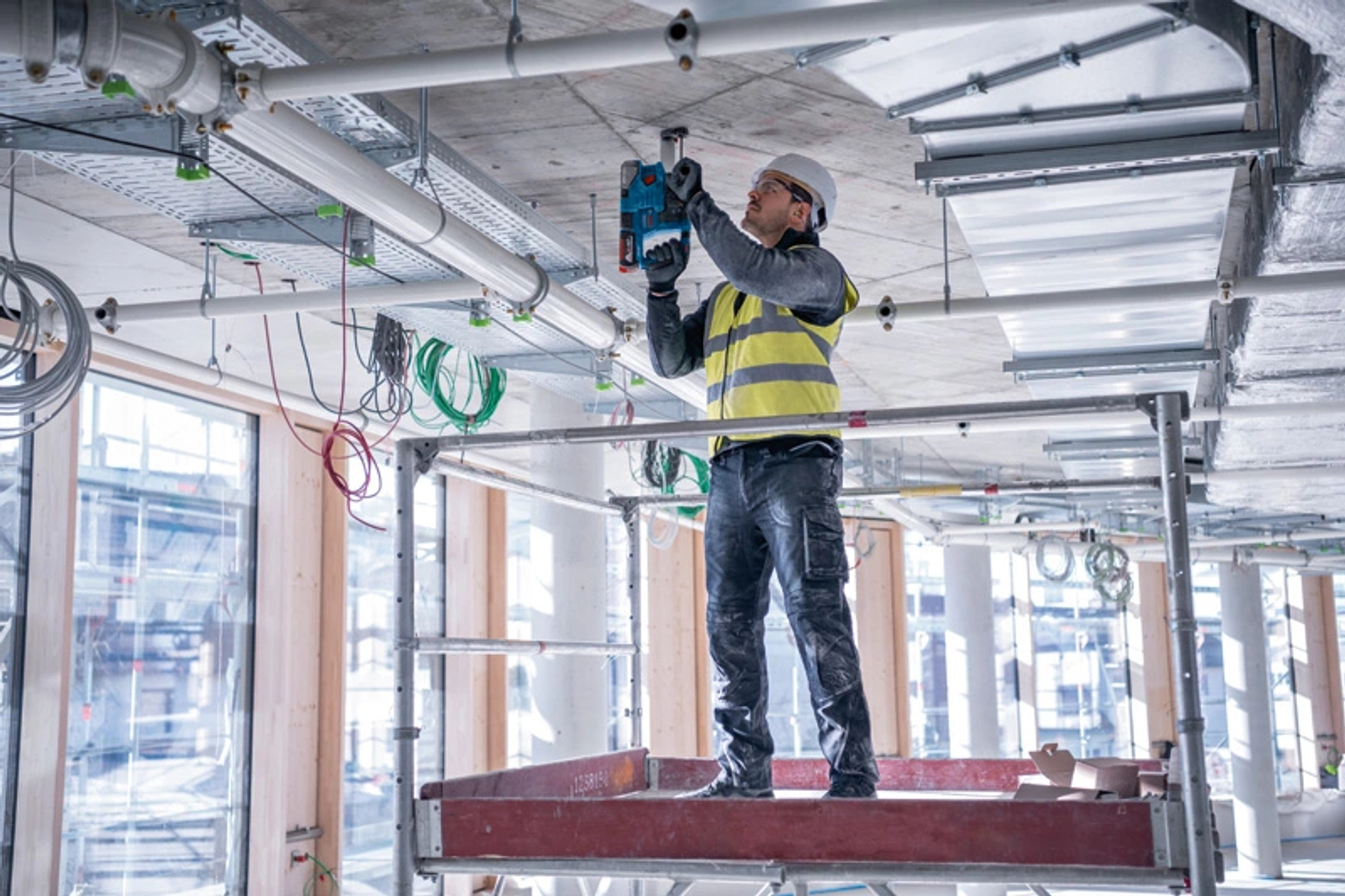 Un trabajador en un andamio usando un taladro en un techo con tuberías y cables. Obra en construcción.