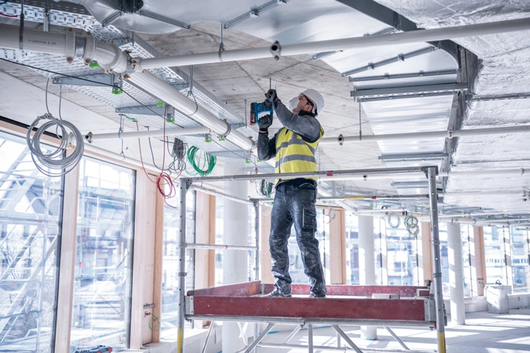 Un trabajador de la construcción en un andamio taladra un techo. La escena es en interiores.