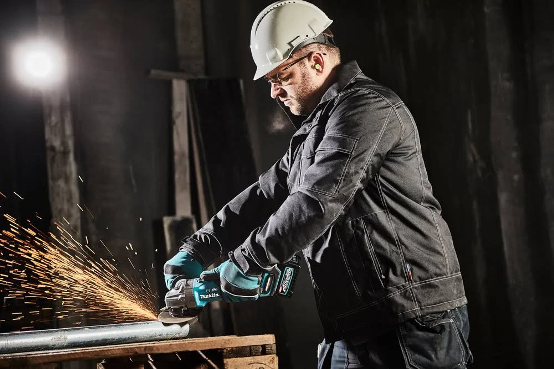 Mężczyzna w białym kasku i okularach ochronnych szlifuje metal, lecą iskry. Szlifierka Makita jest turkusowa.