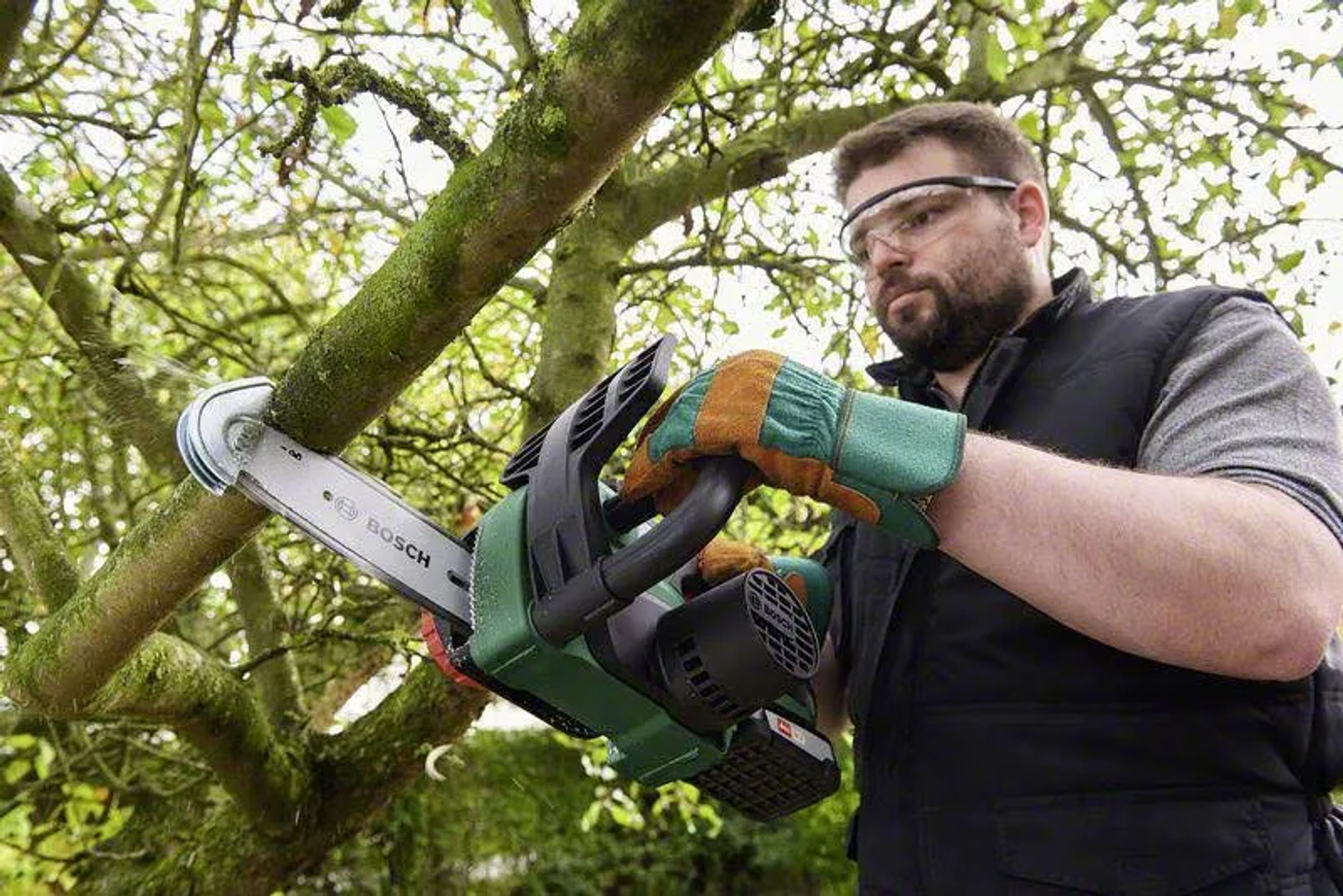 Mężczyzna używa piły łańcuchowej Bosch do cięcia gałęzi. Nosi okulary ochronne i rękawice.