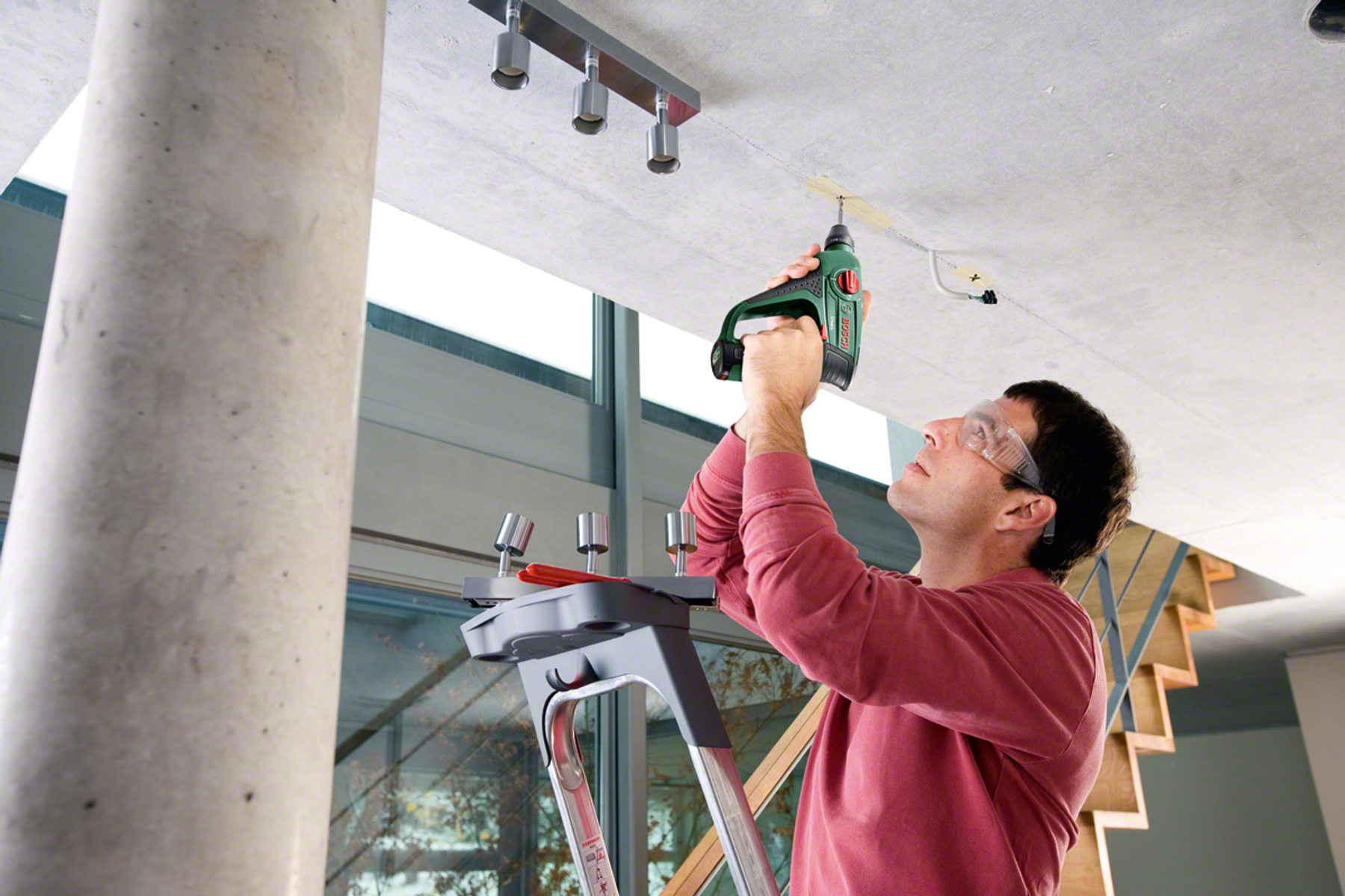 Mężczyzna wierci otwór w białym suficie. Stoi na drabinie, ma na sobie okulary ochronne.