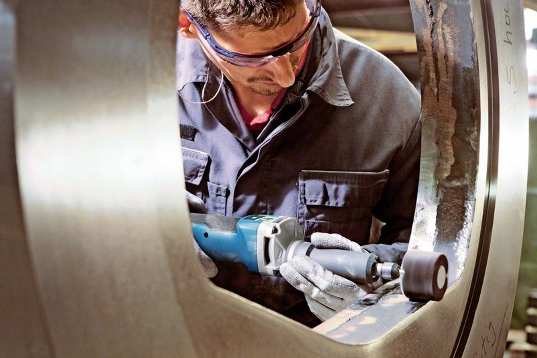 Un homme meule une structure métallique avec un outil électrique. Il porte des lunettes et des gants de sécurité.