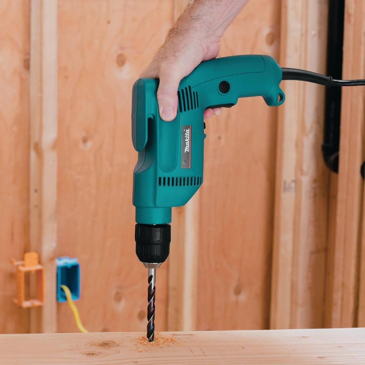 Eine Person bohrt mit einer türkisfarbenen Bohrmaschine ein Loch in Holz. Die Wand ist Holz.