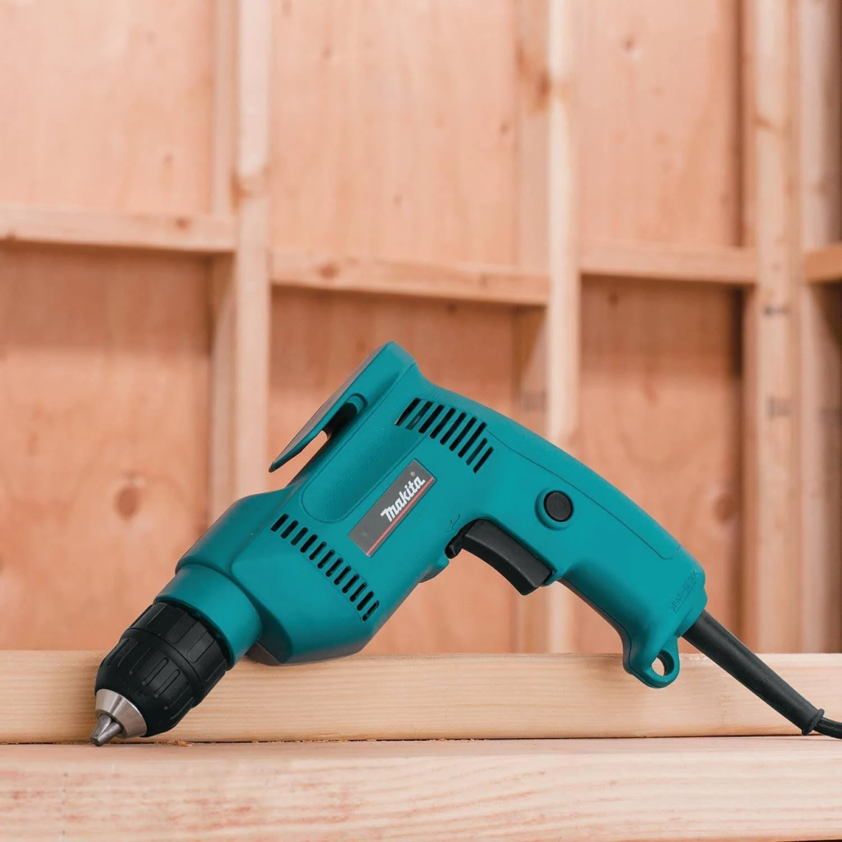 Türkisfarbene Makita-Bohrmaschine auf Holz, abgewinkelt. Schwarzer Griff, silberne Spitze. Holz-Wand-Hintergrund.