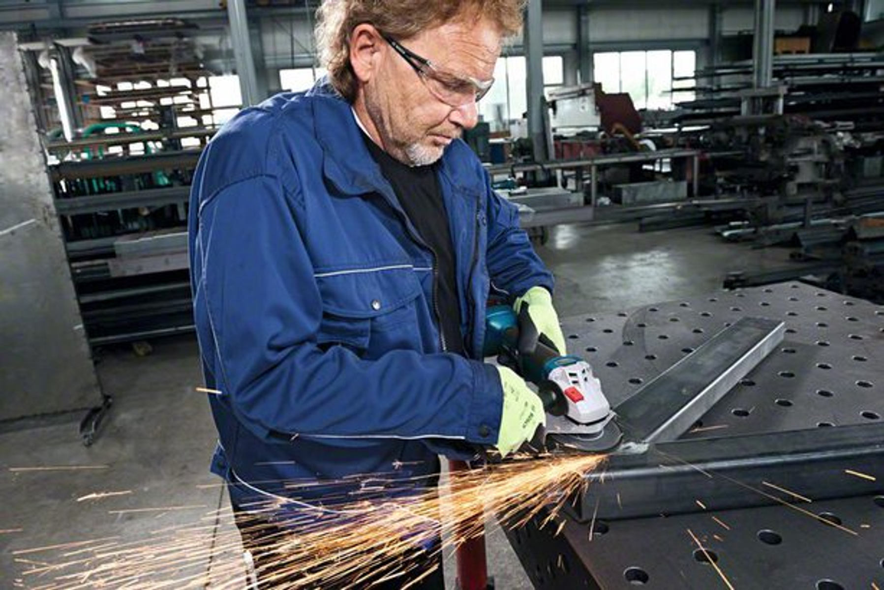 Mężczyzna szlifuje metal. Lecą iskry. Noszenie okularów. Niebieska kurtka. Metalowa powierzchnia.