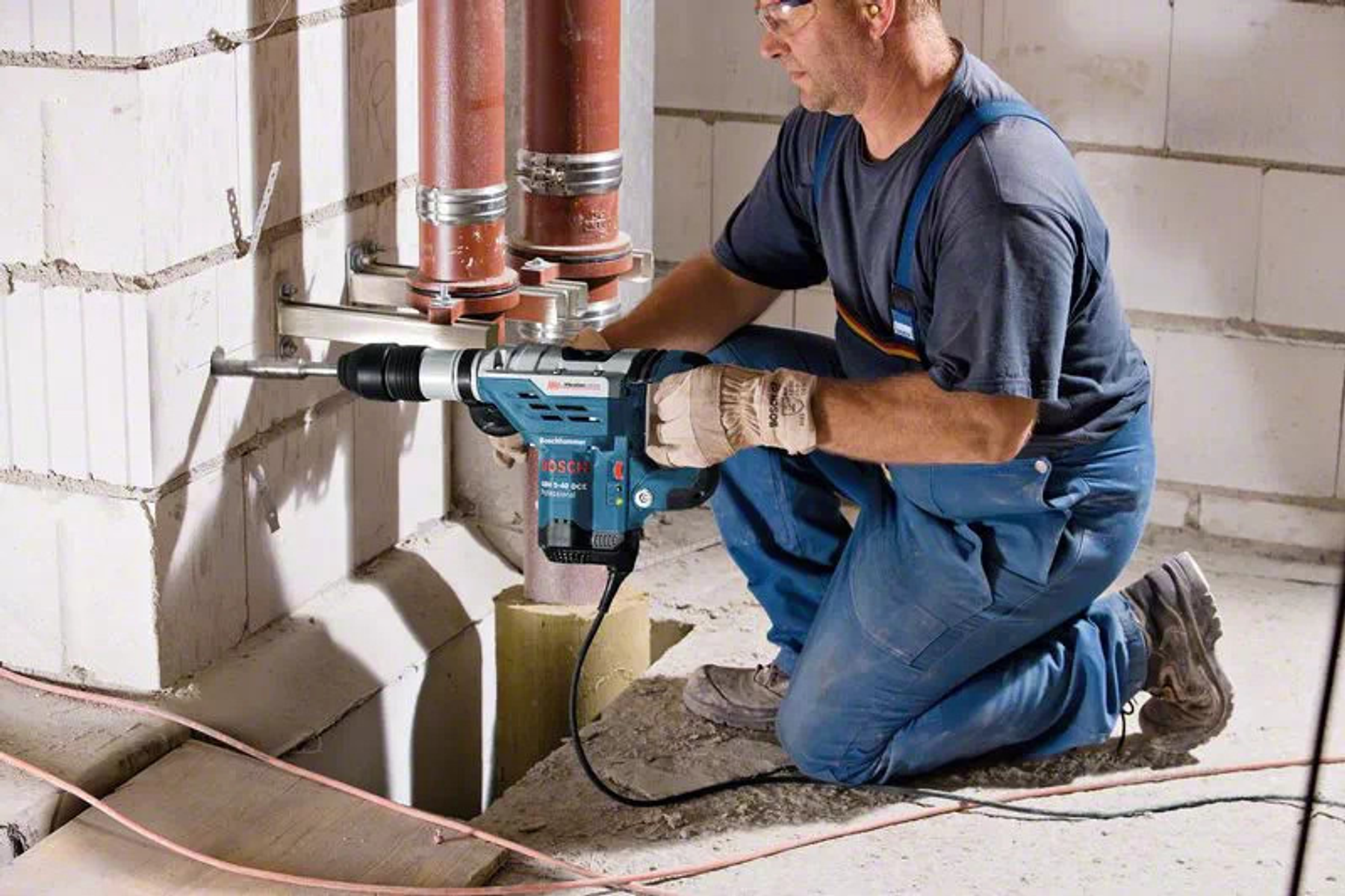Osoba wiercąca w jasnej ceglanej ścianie wiertarką Bosch. Widoczne są rury.