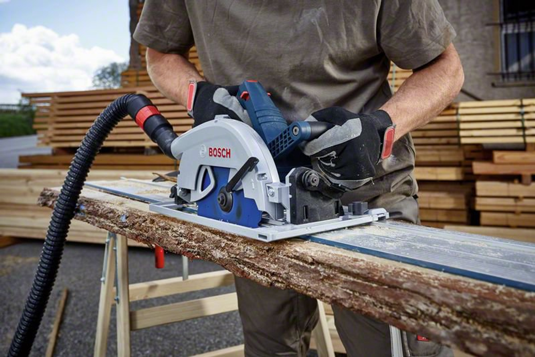 Un hombre usa una sierra circular Bosch para cortar una tabla de madera. La sierra es azul y gris.