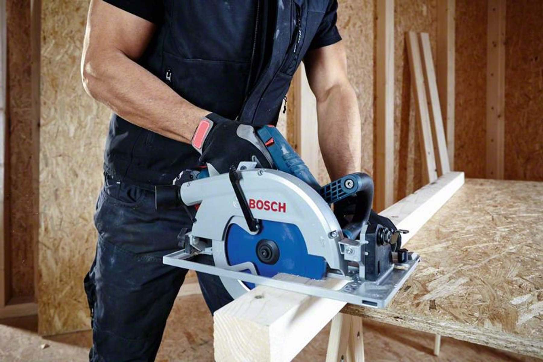 Una persona usa una sierra circular para cortar una tabla de madera. La sierra es azul y gris.