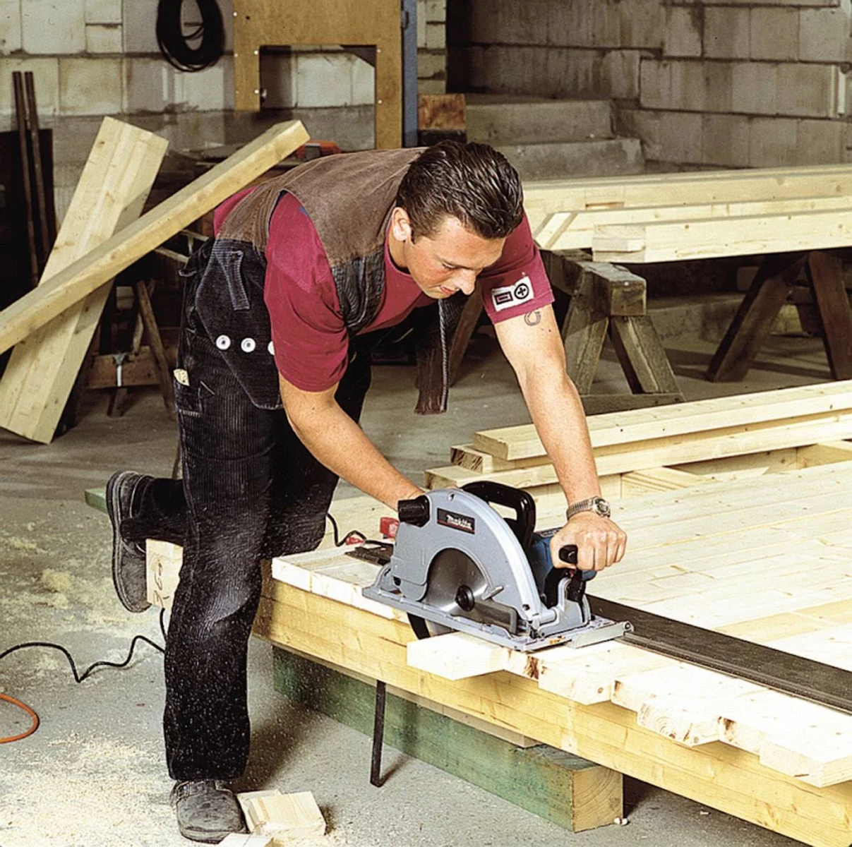 Mężczyzna używa piły tarczowej Makita do cięcia drewna. Ma czerwoną koszulę i czarne spodnie, na drewnianej powierzchni.