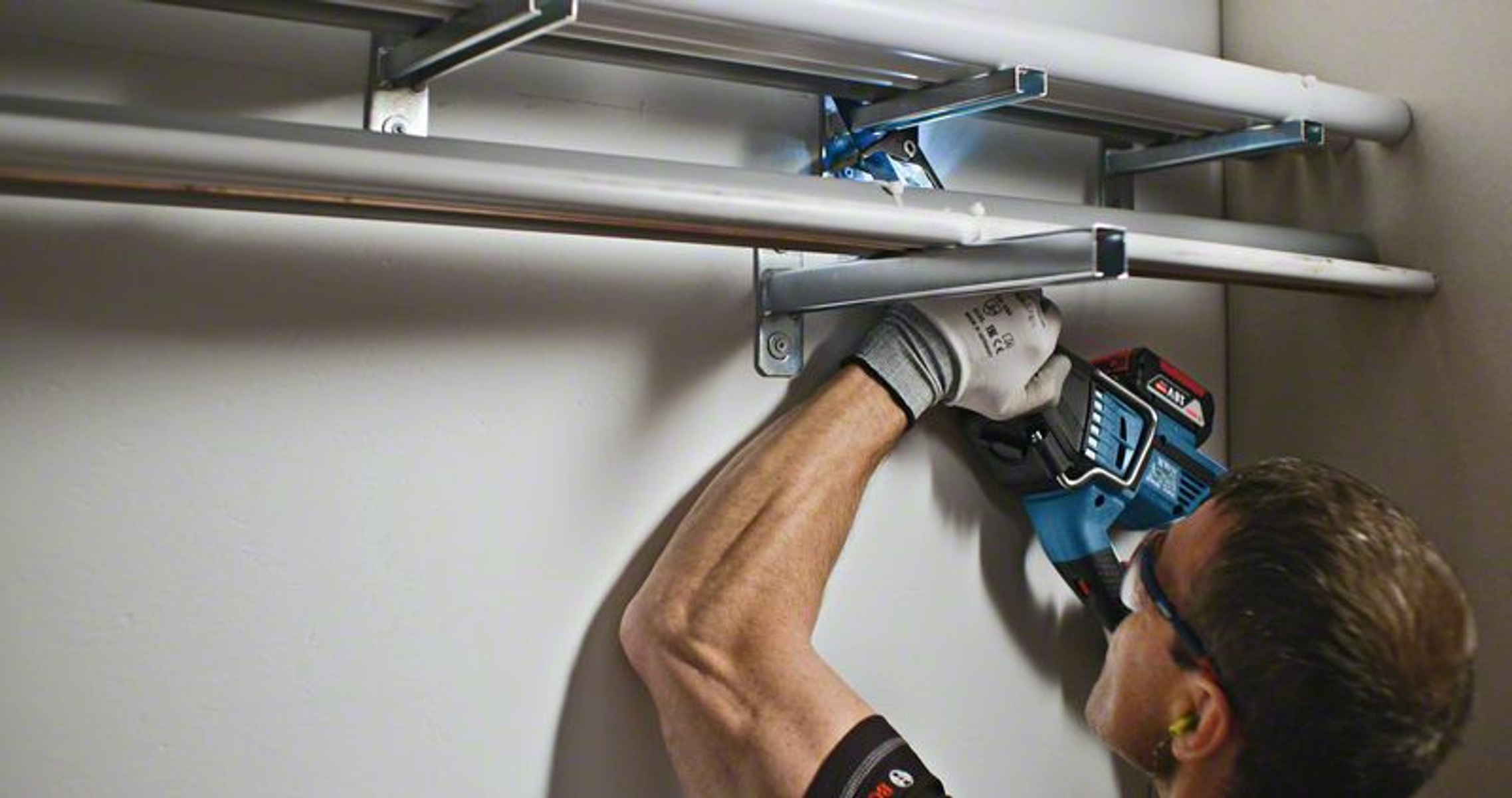 Eine Person, die ein Elektrowerkzeug verwendet, um eine Metallschiene an einer grauen Wand zu installieren.