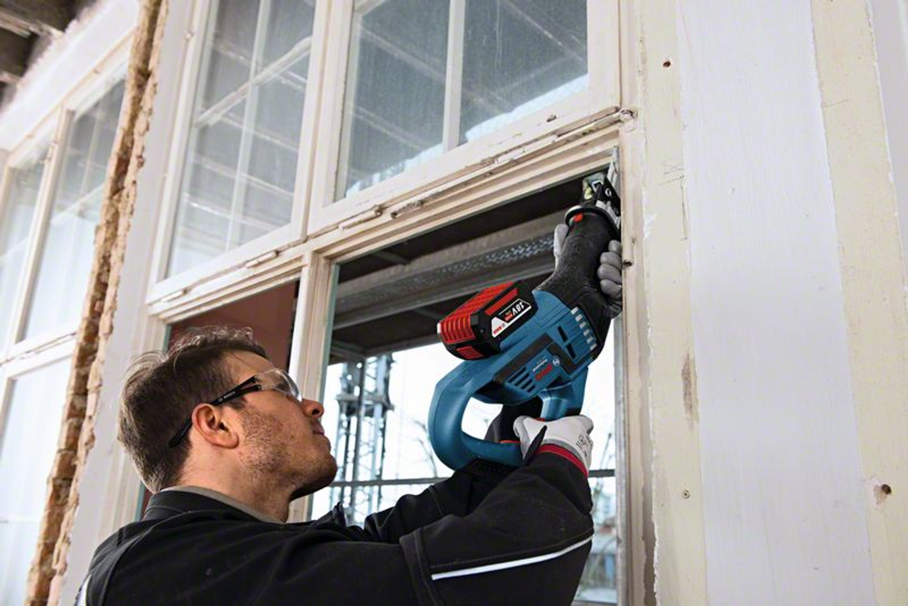 Mann benutzt eine blau-rote Bosch Säbelsäge an einem Fensterrahmen. Brille und Handschuhe sichtbar.