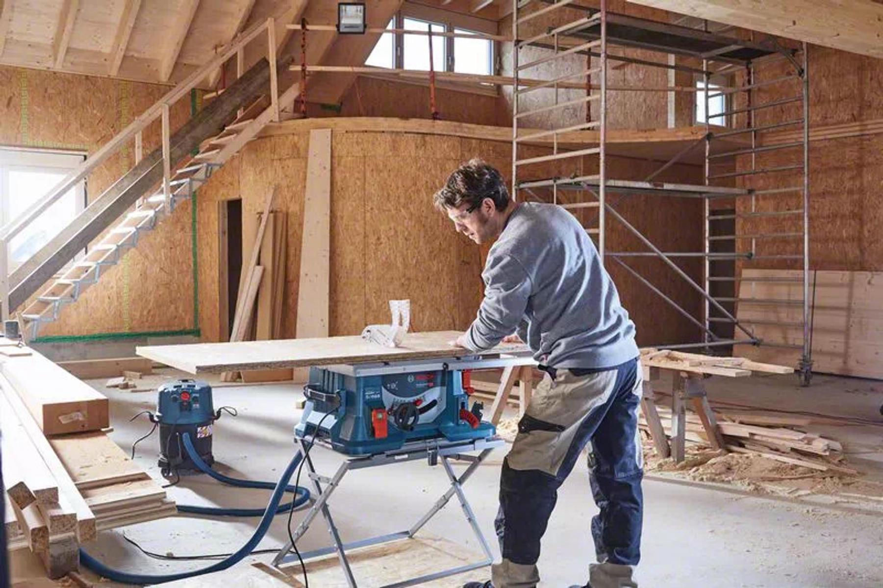 Uomo usa sega Bosch. Segando legno su un tavolo. Interno con ponteggi, scale e legno.