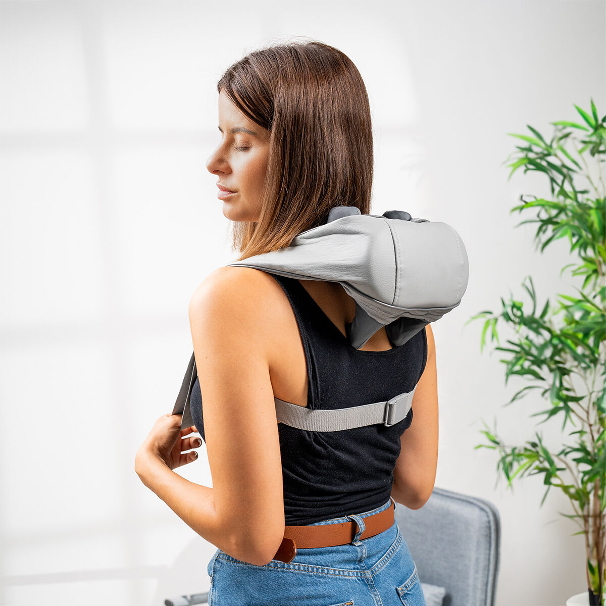 Mujer usando masajeador de cuello y hombros. Dispositivo gris, camiseta negra, jeans azules. Planta al fondo.