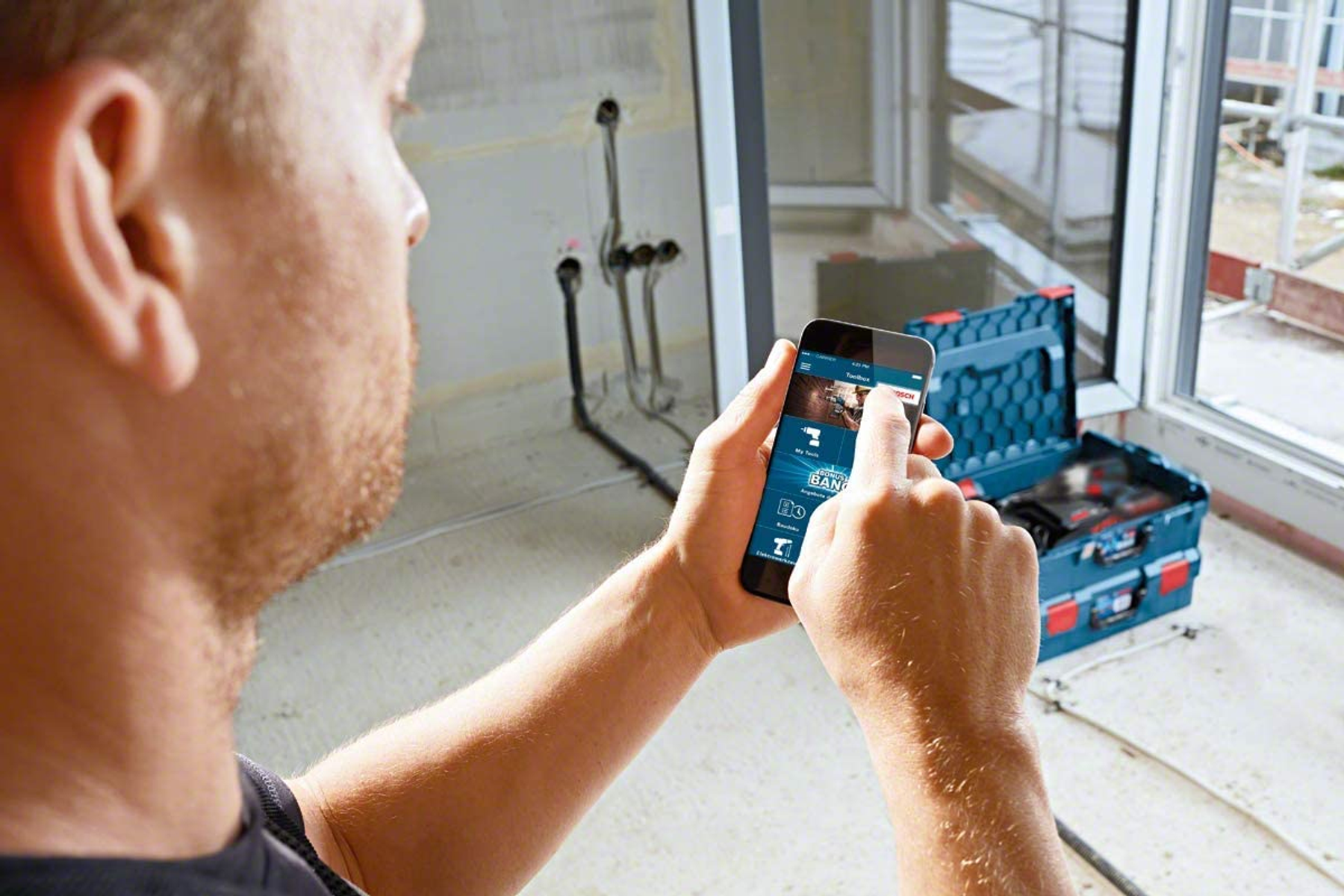 Mann, der ein Smartphone zur Steuerung von Elektrowerkzeugen verwendet. Im Hintergrund eine Baustelle.