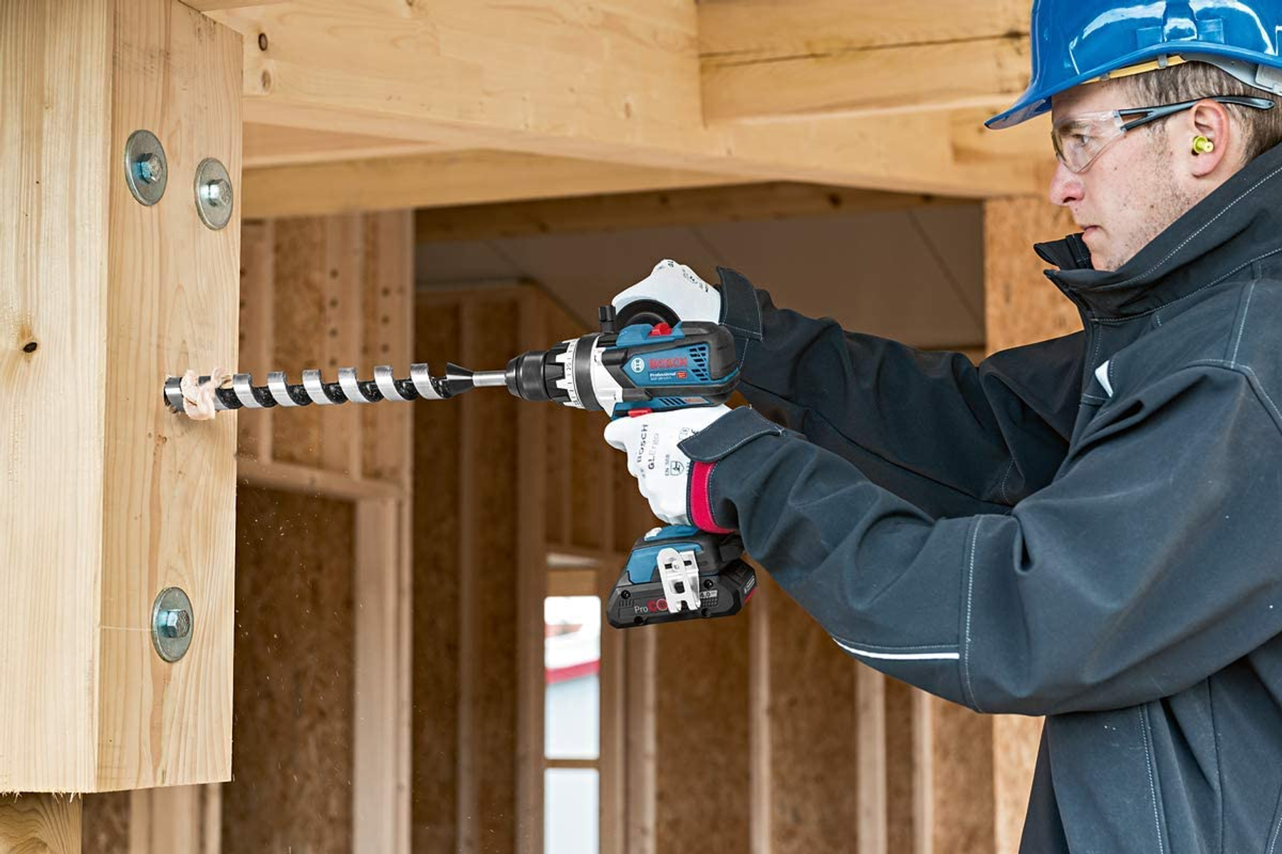 Ein Mann bohrt mit einem Bosch-Bohrer in einen Holzpfosten. Der Hintergrund ist eine Baustelle.