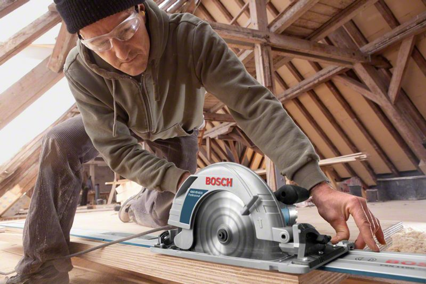Ein Mann benutzt eine Bosch-Kreissäge auf einer Holzplanke. Innenräume mit Holzbalken.