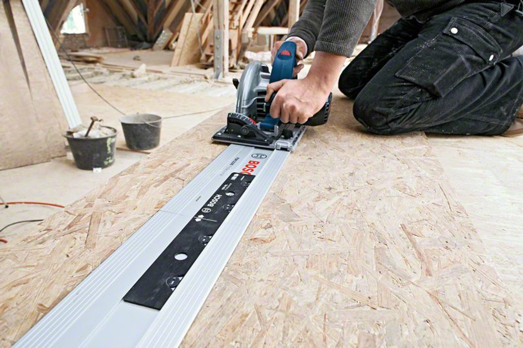 Eine Person schneidet mit einer Bosch-Kreissäge eine Holzplatte. Säge auf einer Führungsschiene. Hintergrund: Baustelle.
