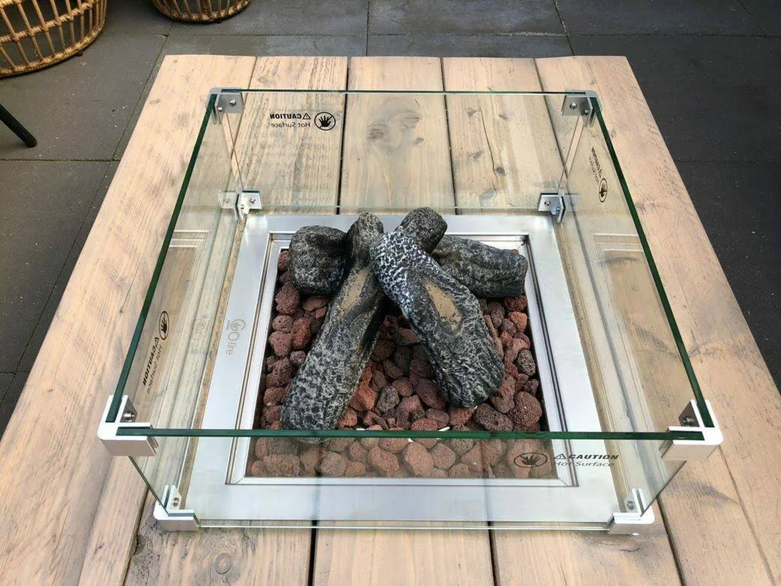 Una fogata cuadrada con vidrio, piedras decorativas y troncos falsos sobre una superficie de madera.