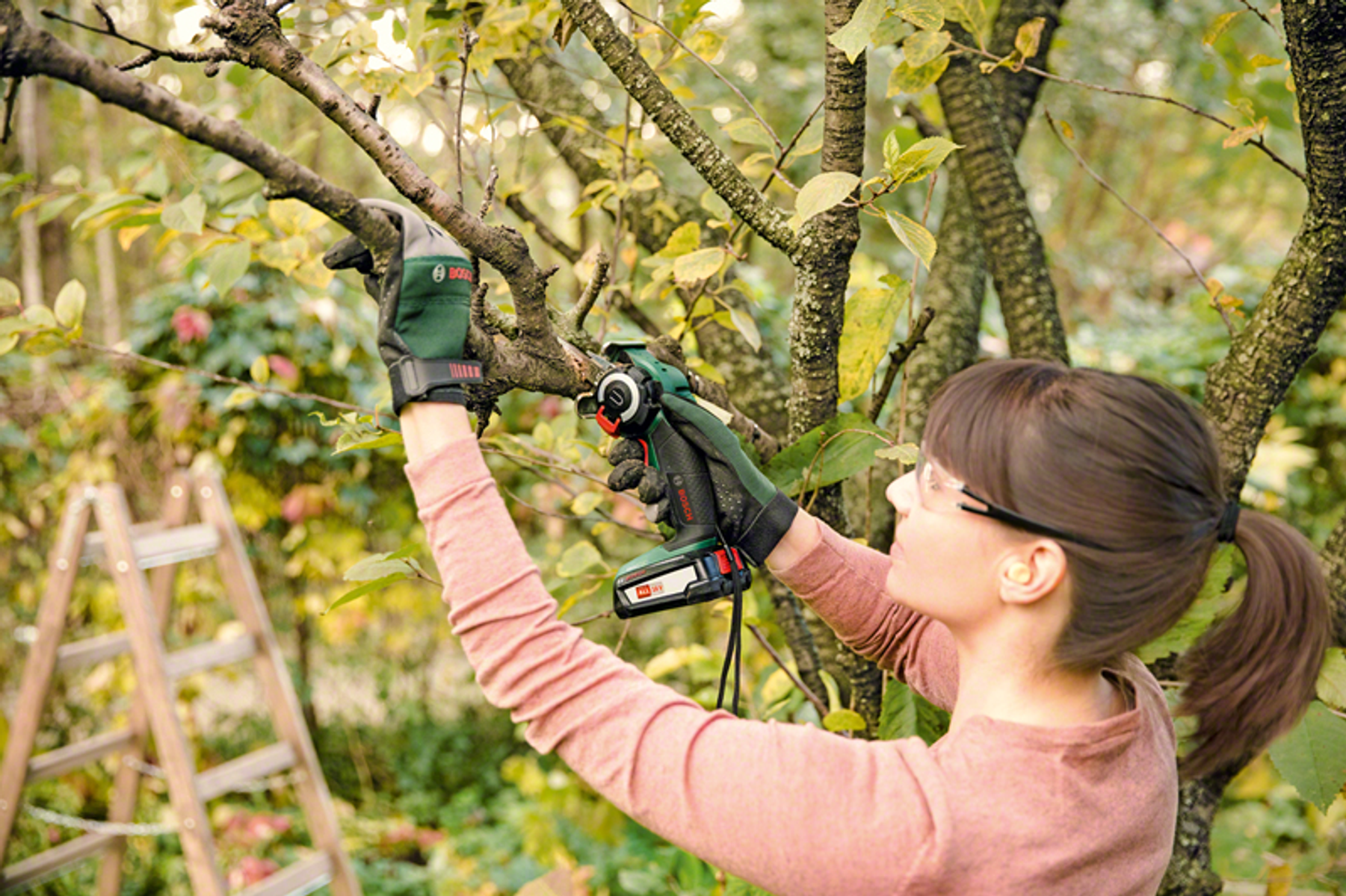 Kobieta używa piły akumulatorowej Bosch do przycinania gałęzi. Nosi okulary i rękawice.