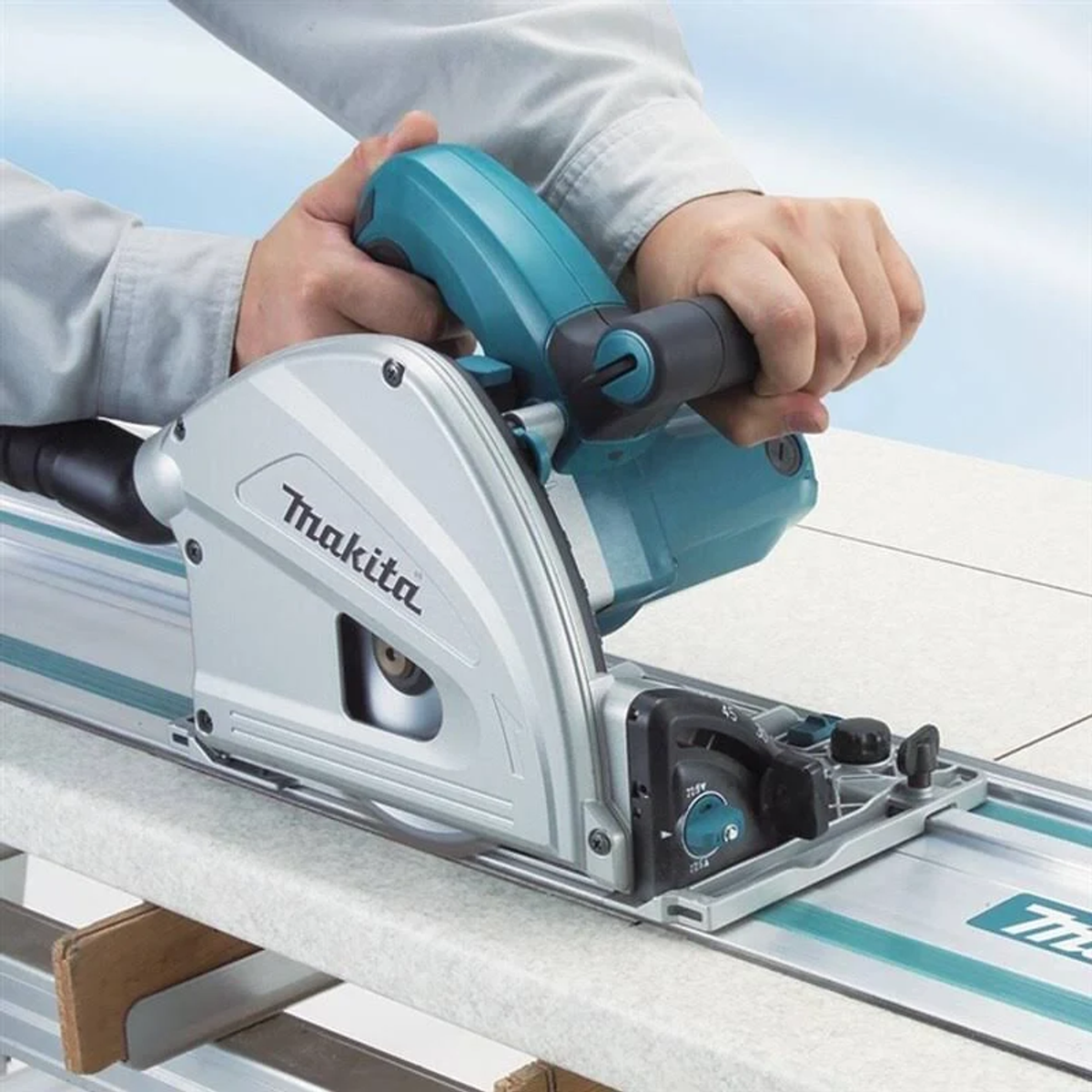 Mains utilisant une scie sur rail Makita pour couper une table blanche. Scie bleue et grise sur une surface blanche.