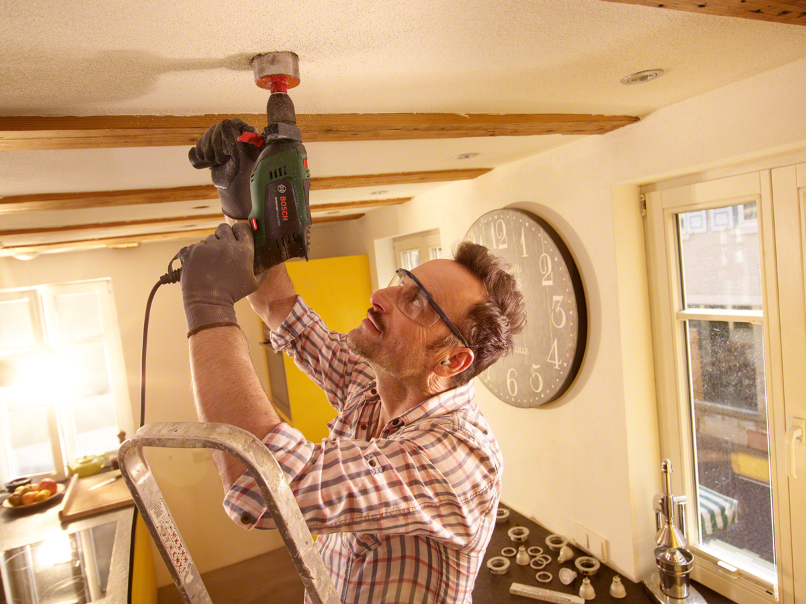 Mężczyzna na drabinie wiercący w suficie. Ma okulary ochronne i rękawice. W tle kuchnia.