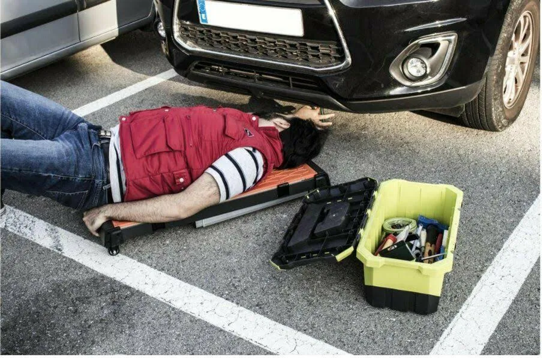 Persona tumbada debajo de un coche, con herramientas en un contenedor amarillo y caja.