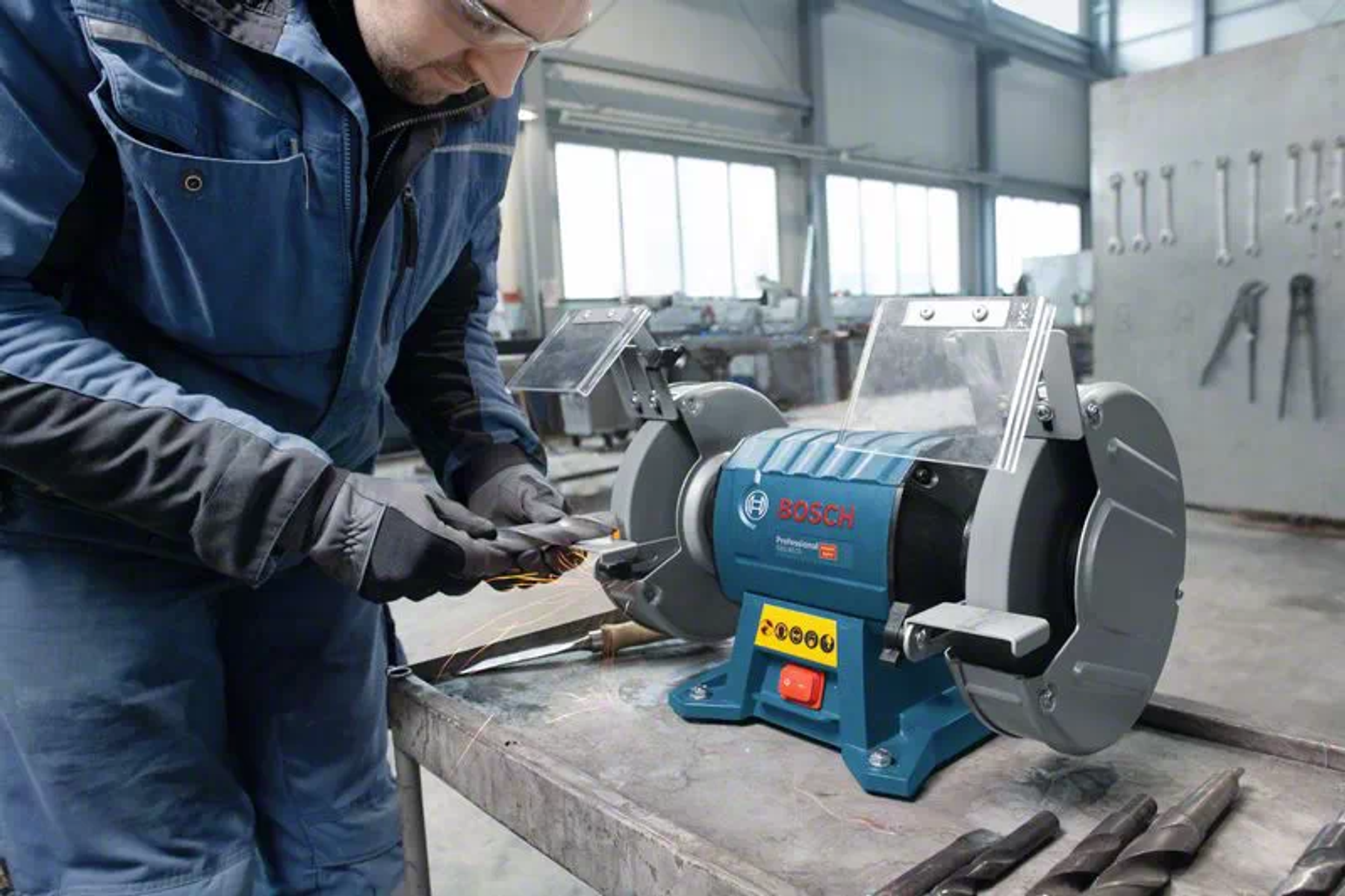 Bir adam, metal bir masa üzerinde Bosch taşlama makinesi kullanıyor, güvenlik gözlüğü ve eldiven takıyor, iç mekanda.