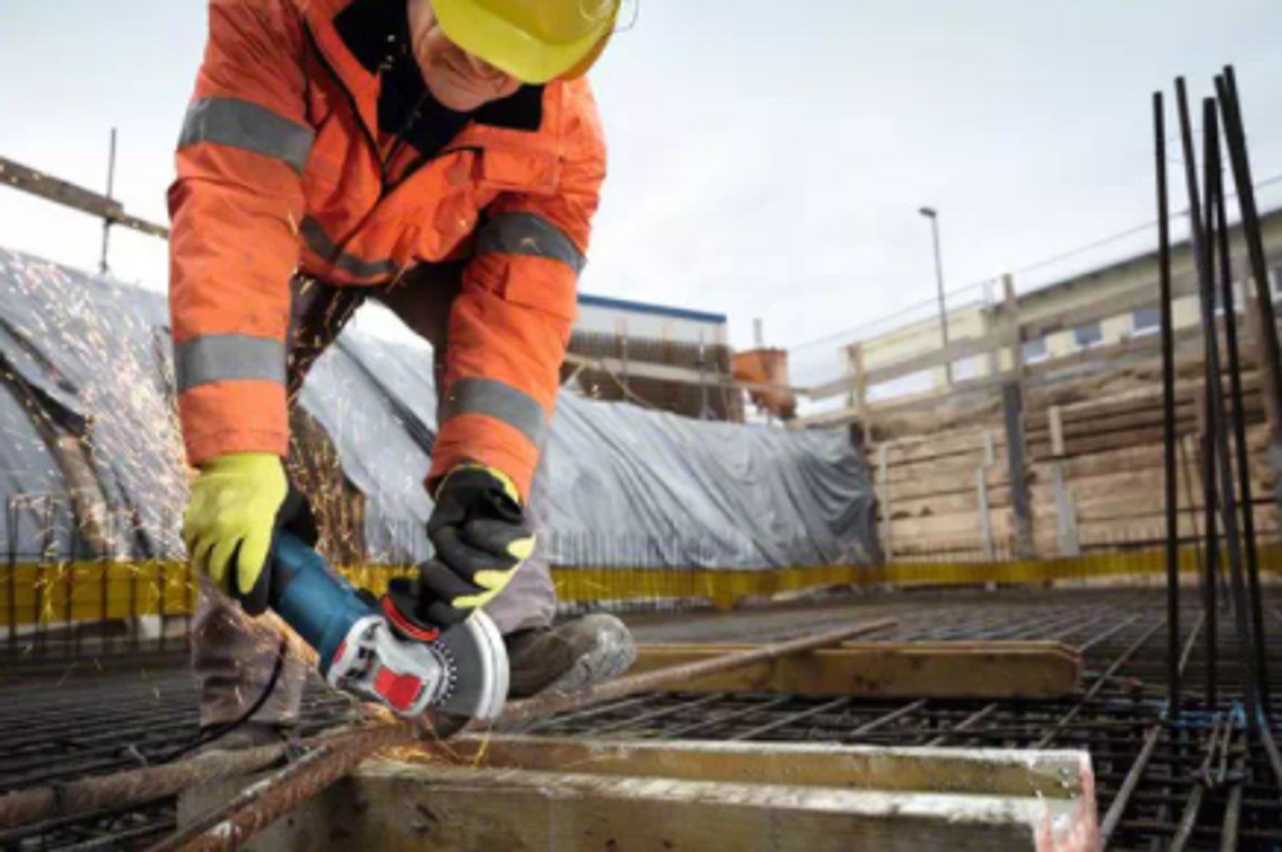 Un trabajador con chaqueta naranja y guantes amarillos usando una amoladora en una barra de refuerzo metálica.