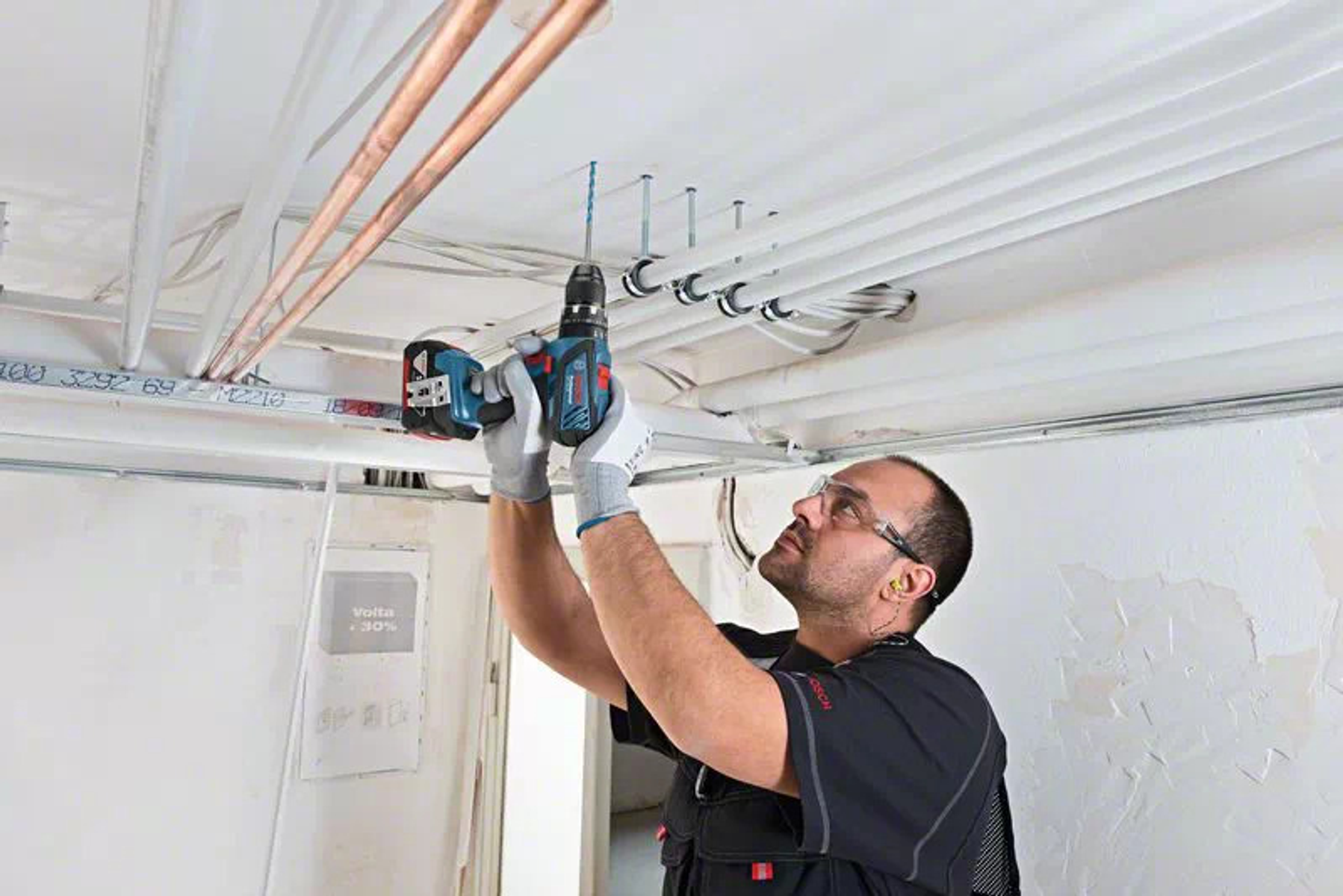 Mężczyzna w rękawicach wierci w suficie niebiesko-czarną wiertarką Bosch, otoczony rurami.