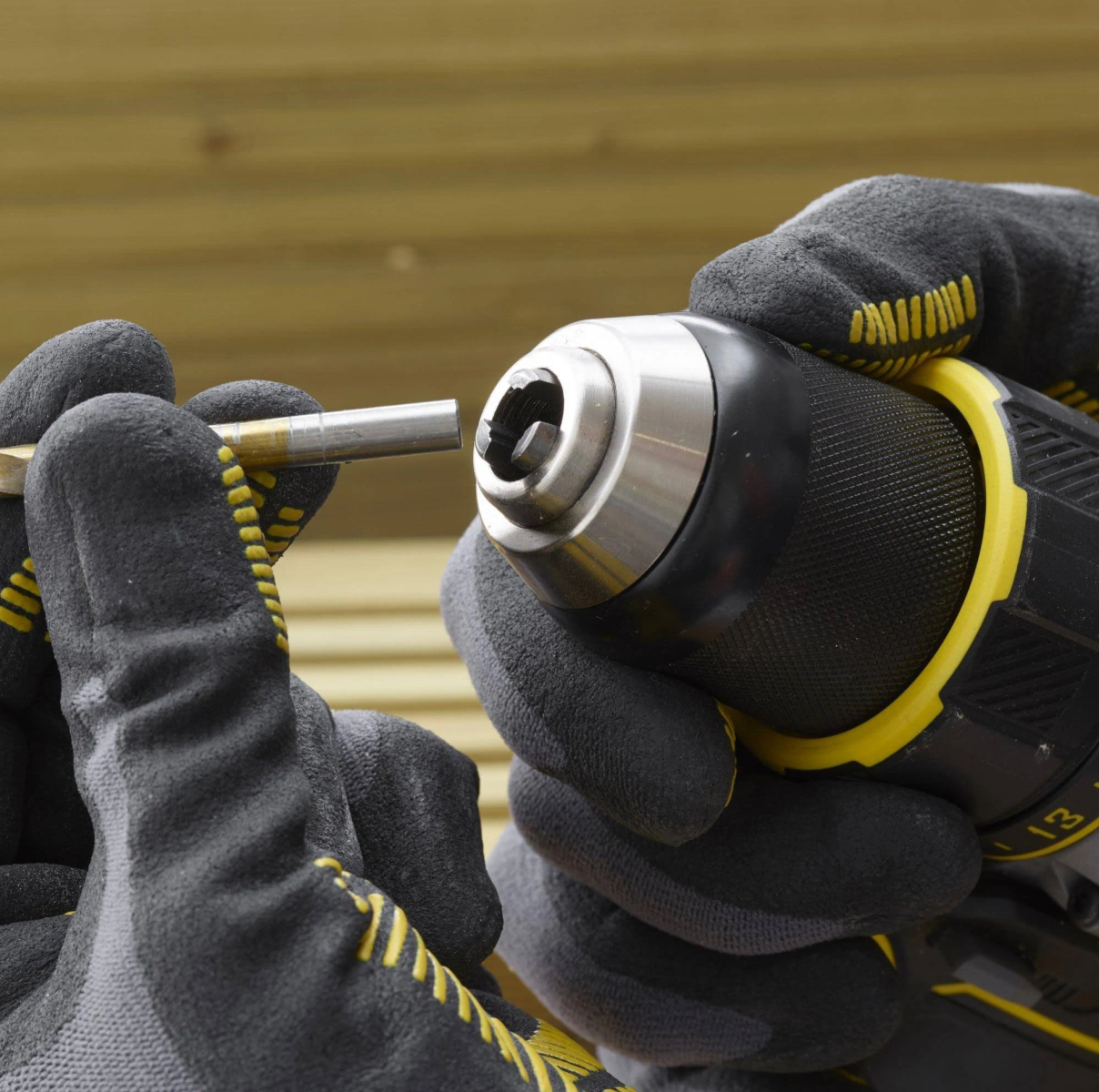 Manos con guantes insertando una broca en un taladro. Colores amarillo y negro, fondo de madera.