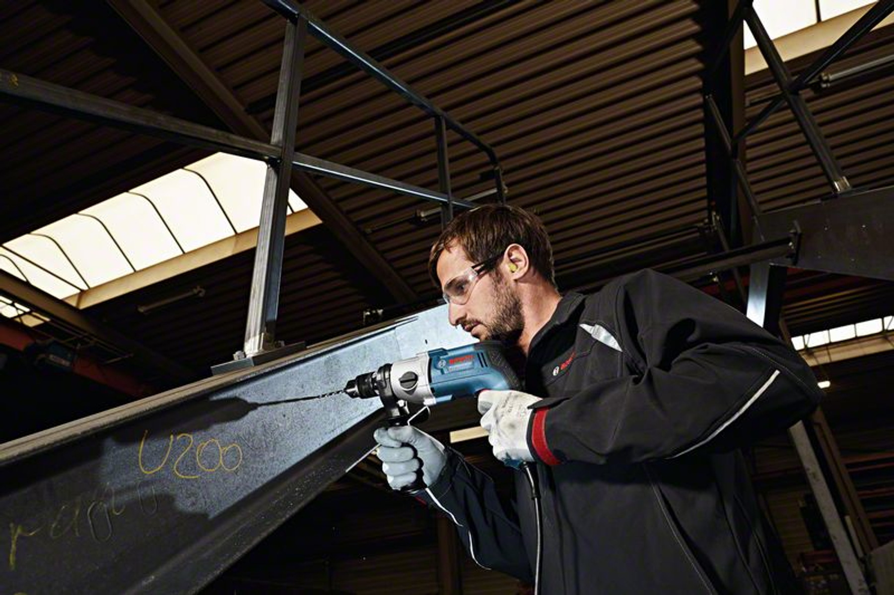 Mężczyzna wierci otwór w metalowej belce, w okularach i zatyczkach.