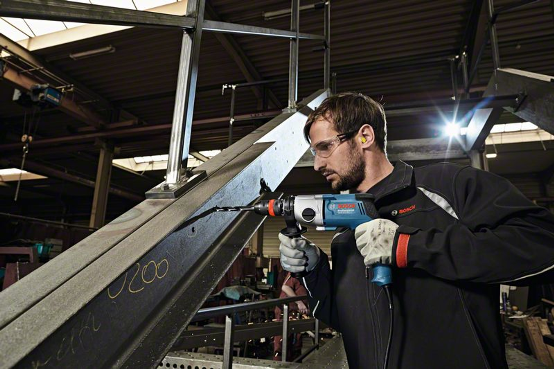 Mężczyzna wierci otwór w metalowej belce. Ma na sobie okulary ochronne i czarną kurtkę, z niebieską wiertarką.