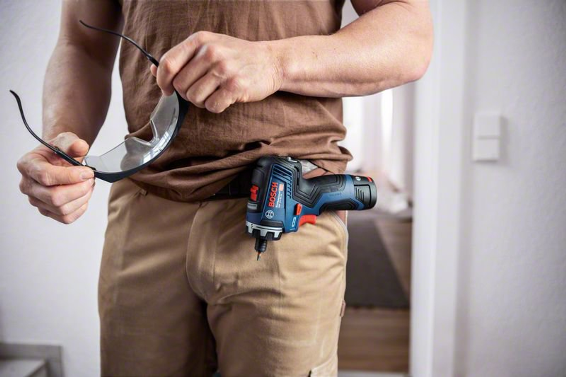 Eine Person hält eine Schutzbrille, während ein Akku-Bohrschrauber an ihrer Hose hängt. Der Bohrschrauber ist schwarz, blau und rot.