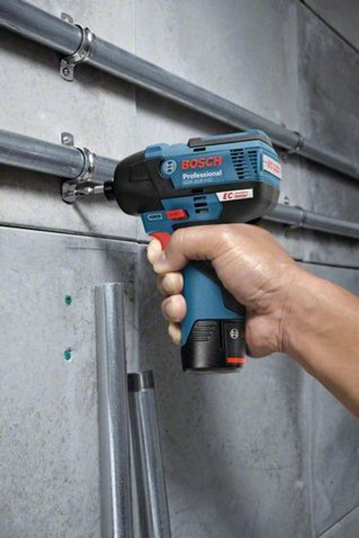 Una mano sosteniendo un atornillador de impacto Bosch azul y negro, taladrando una pared gris con tuberías metálicas.