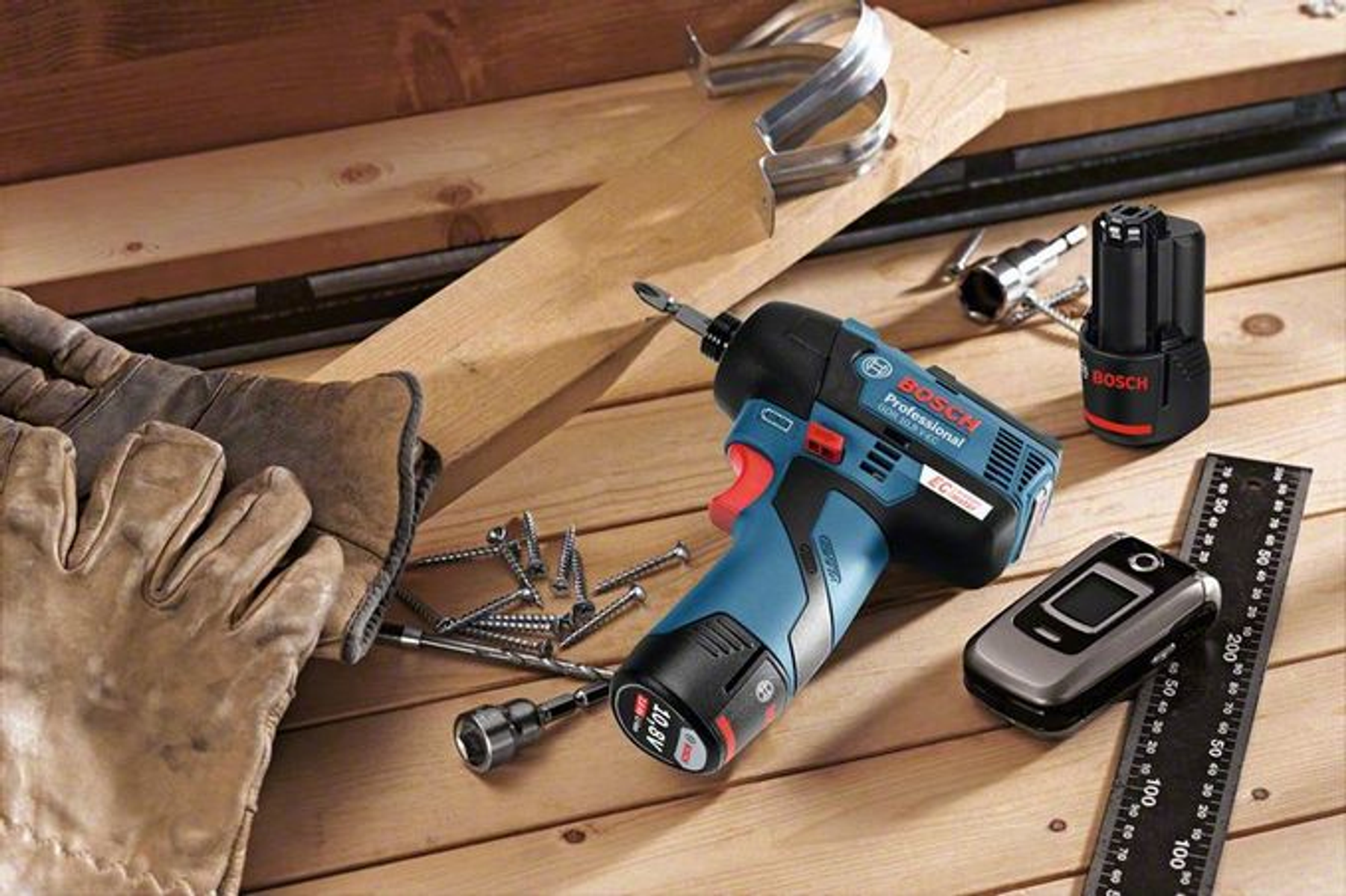Un taladro Bosch inalámbrico y accesorios en una superficie de madera con guantes y tornillos.