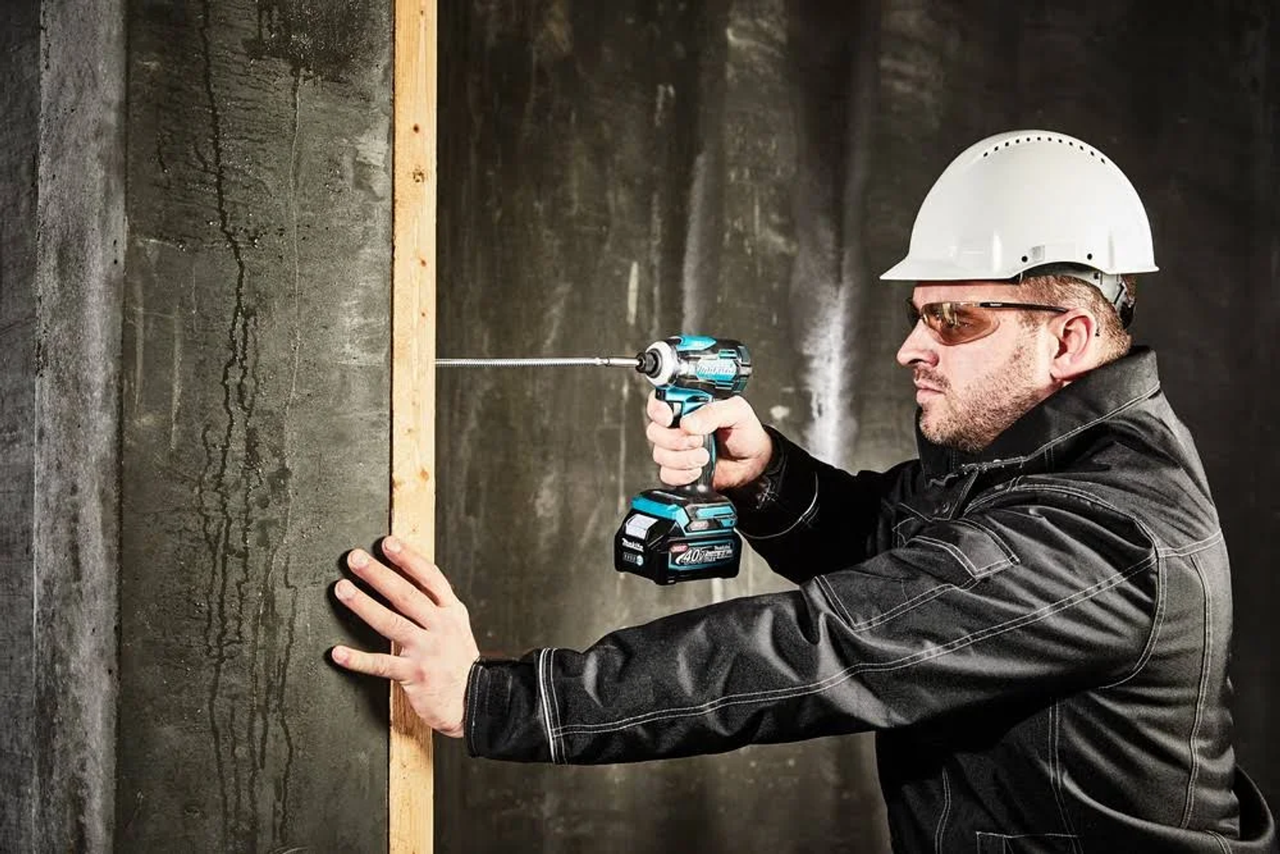 Un uomo usa un trapano turchese e nero per avvitare in un montante a muro. Indossa un casco.