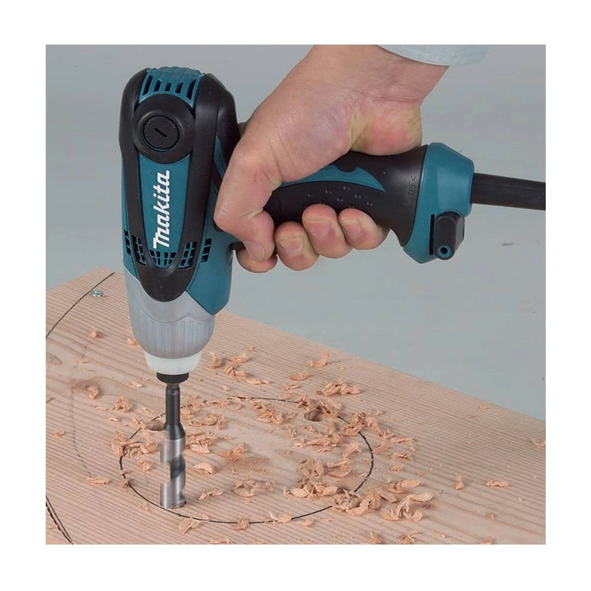Osoba wierci okrągły otwór w drewnie za pomocą wiertarki Makita. Wióry drewniane obecne, białe tło.