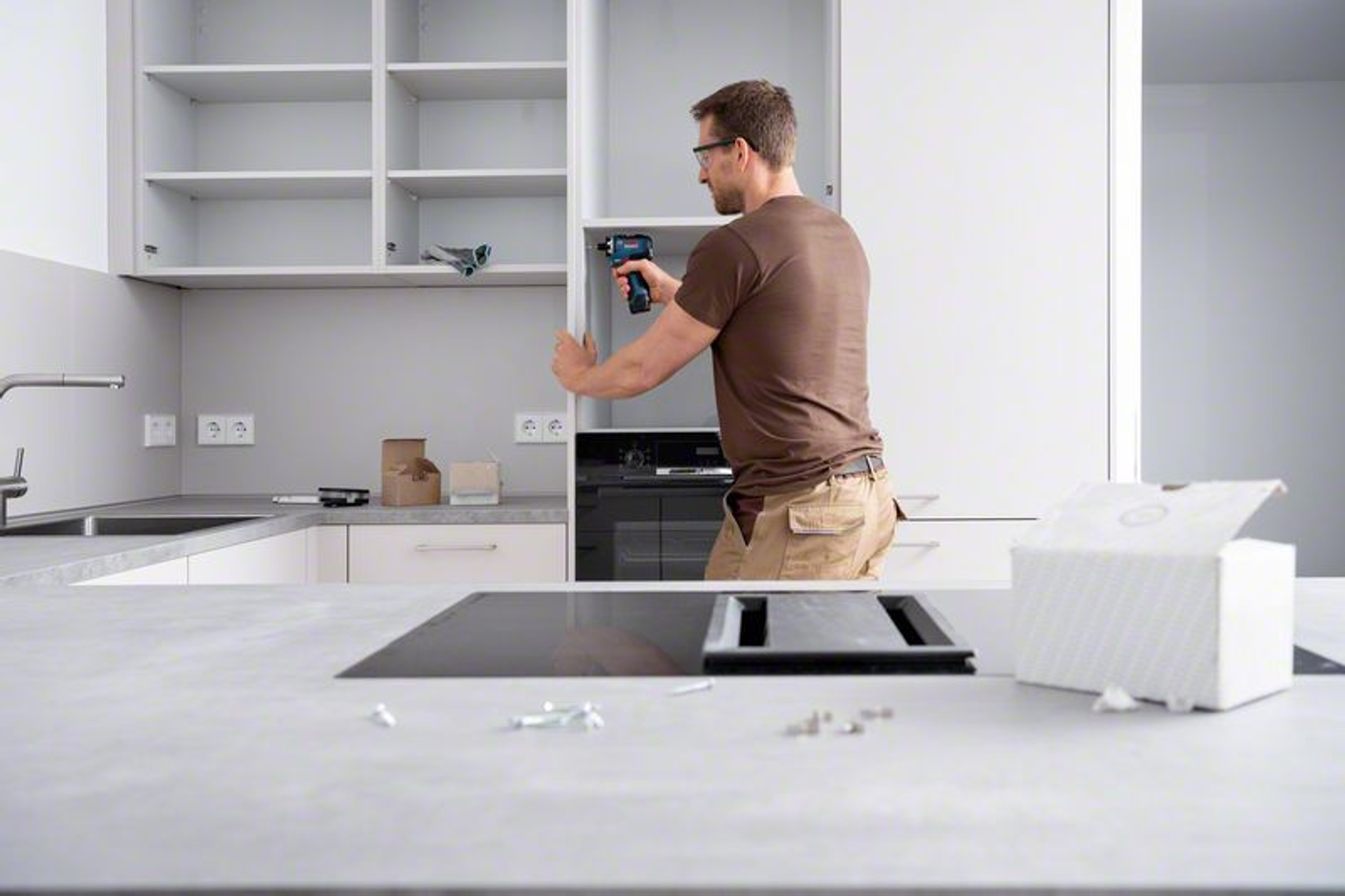 Mężczyzna montuje szafkę kuchenną wiertarką. Białe szafki, szary blat i czarne urządzenia.