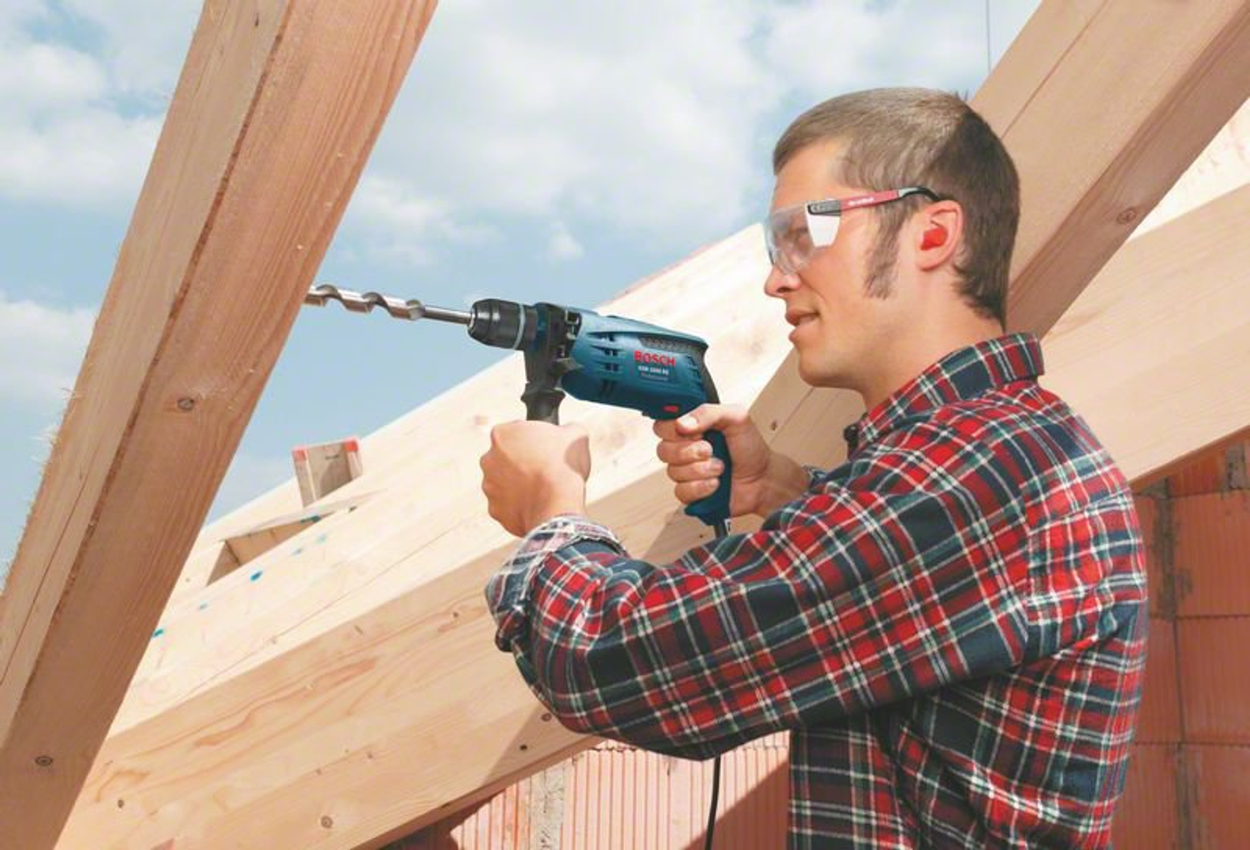 Un uomo sta forando travi di legno con un trapano Bosch, cielo visibile sullo sfondo.