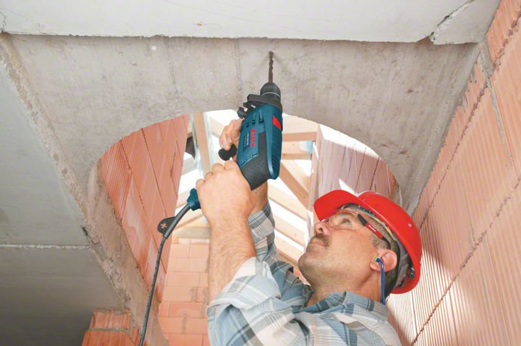 Un uomo con un casco rosso che fora un soffitto di cemento con un trapano blu.