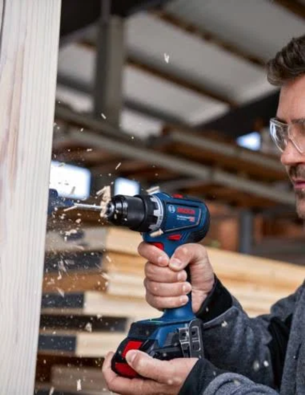 Un hombre con gafas está usando un taladro para perforar una estructura de madera. El taladro es azul y negro.