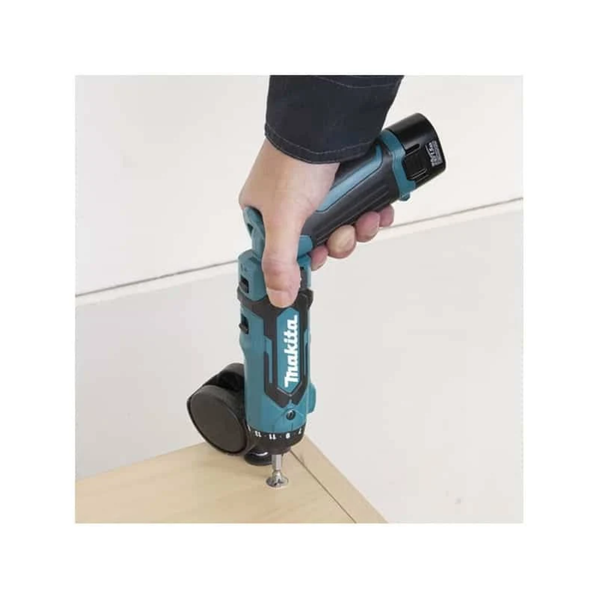 Ręka używa turkusowej wiertarki Makita do wkręcania w drewnianą powierzchnię. Tło to biała ściana.