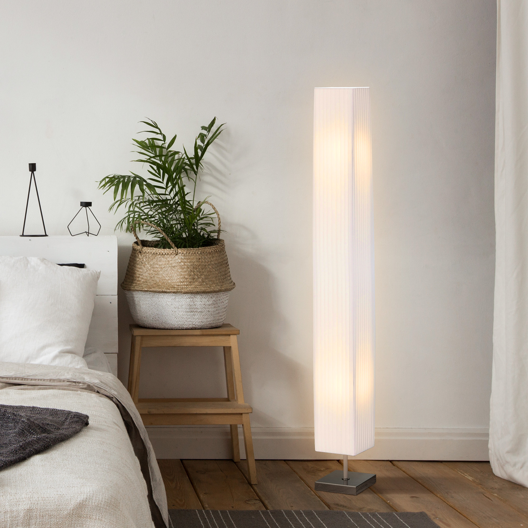 Camera da letto con lampada da terra, letto, pianta su sgabello e tende bianche.