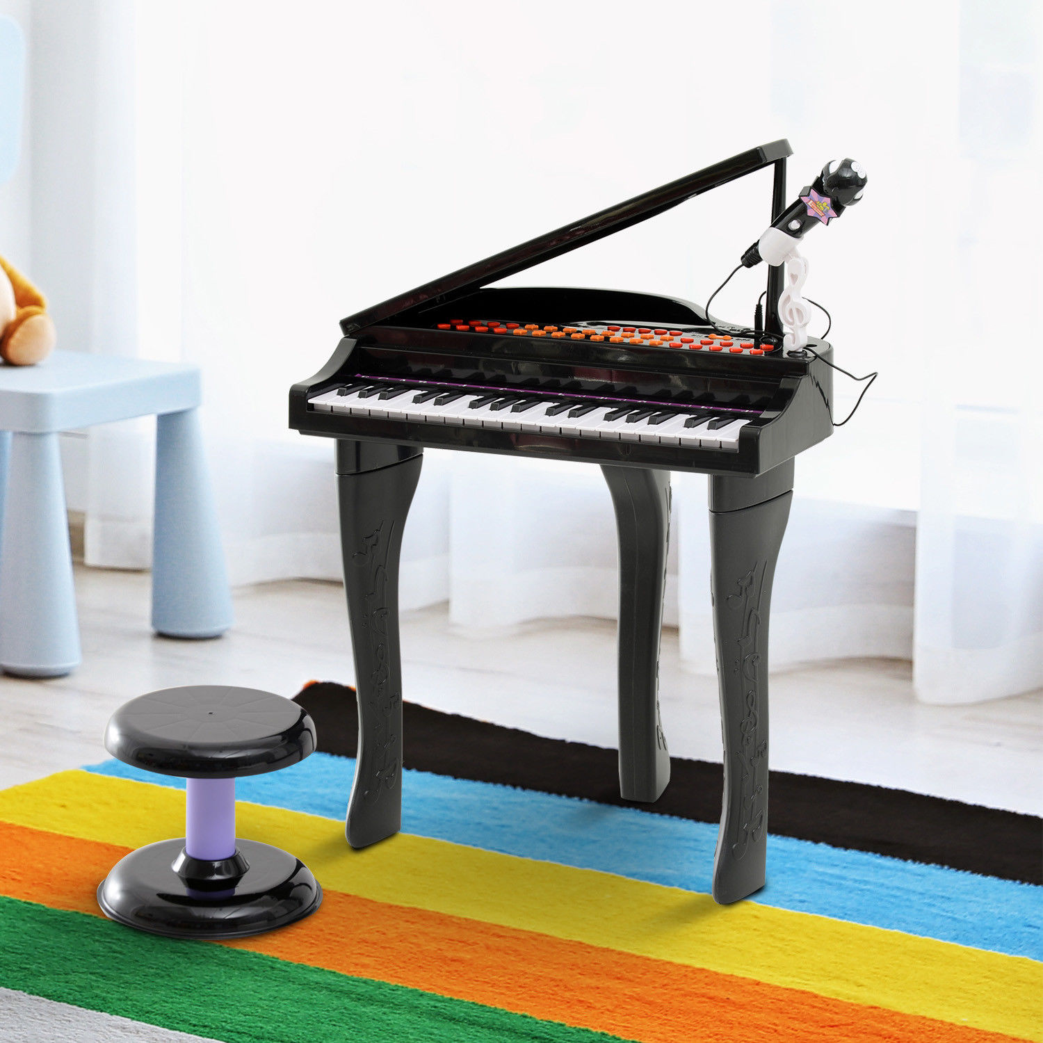 Pianoforte giocattolo nero con microfono, tasti bianchi e sgabello, su un tappeto colorato.