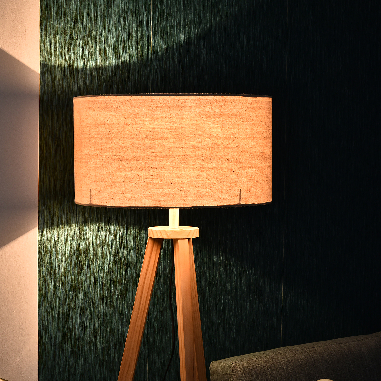 Lampada a treppiede in legno con paralume cilindrico beige contro un muro verde acqua.