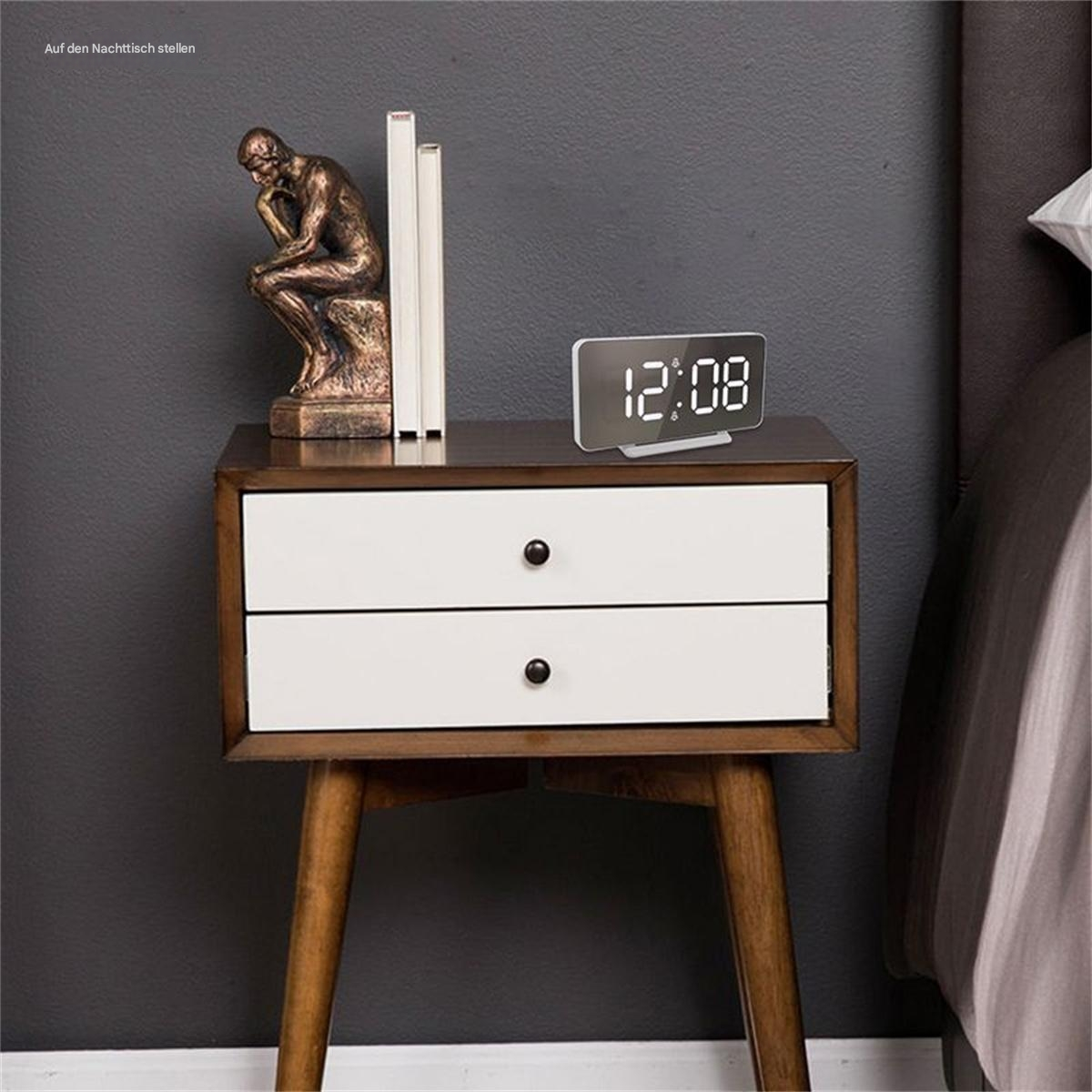 Nachttisch mit Digitaluhr, Büchern und einer Statue. Dunkelgrauer Wandhintergrund.